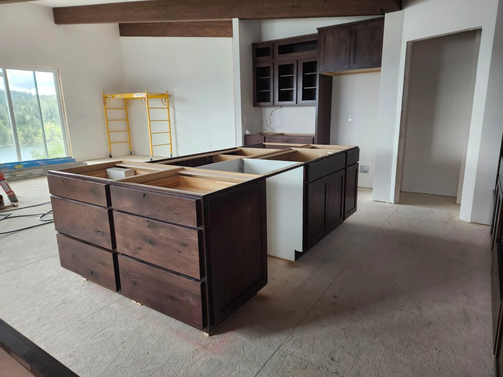 Kitchen under construction with dark wood cabinets, an island with incomplete countertop, a window showing a scenic outdoor view, and construction tools and scaffolding present.
