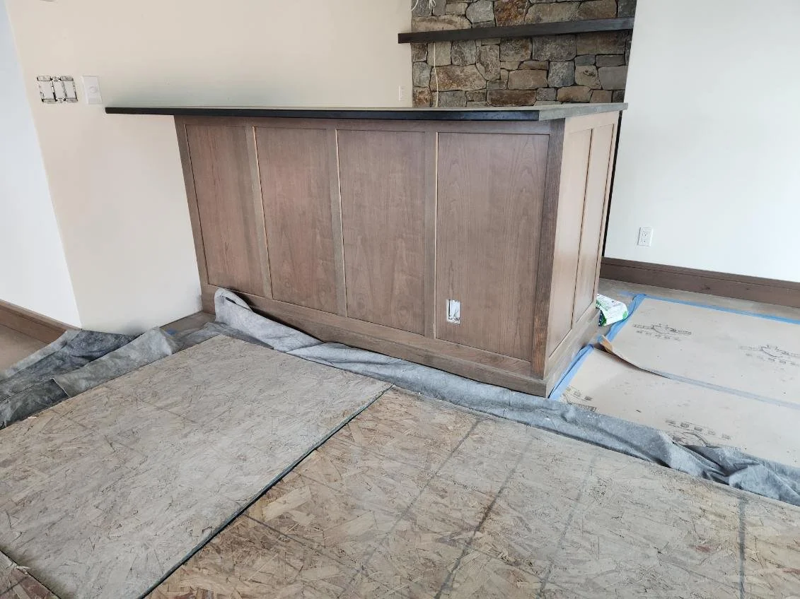 Interior under renovation showing a wooden bar or counter with a gray countertop, a stone wall in the background, and flooring protected with plywood and plastic sheeting.