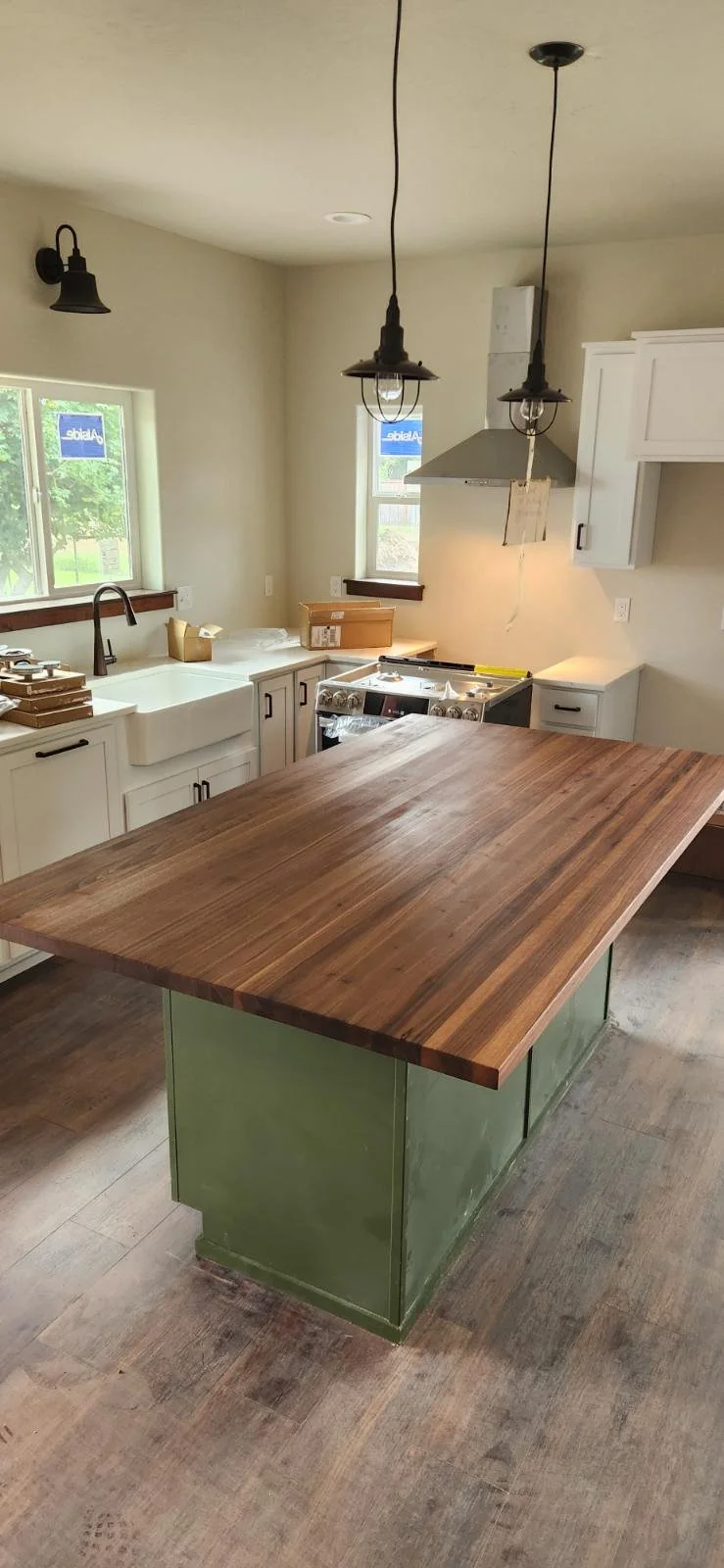 Coeur d'Alene Modern kitchen with white cabinets, black handles, a farmhouse sink, a wooden kitchen island, industrial pendant lights, a stove with a range hood, and two windows showing outside greenery.