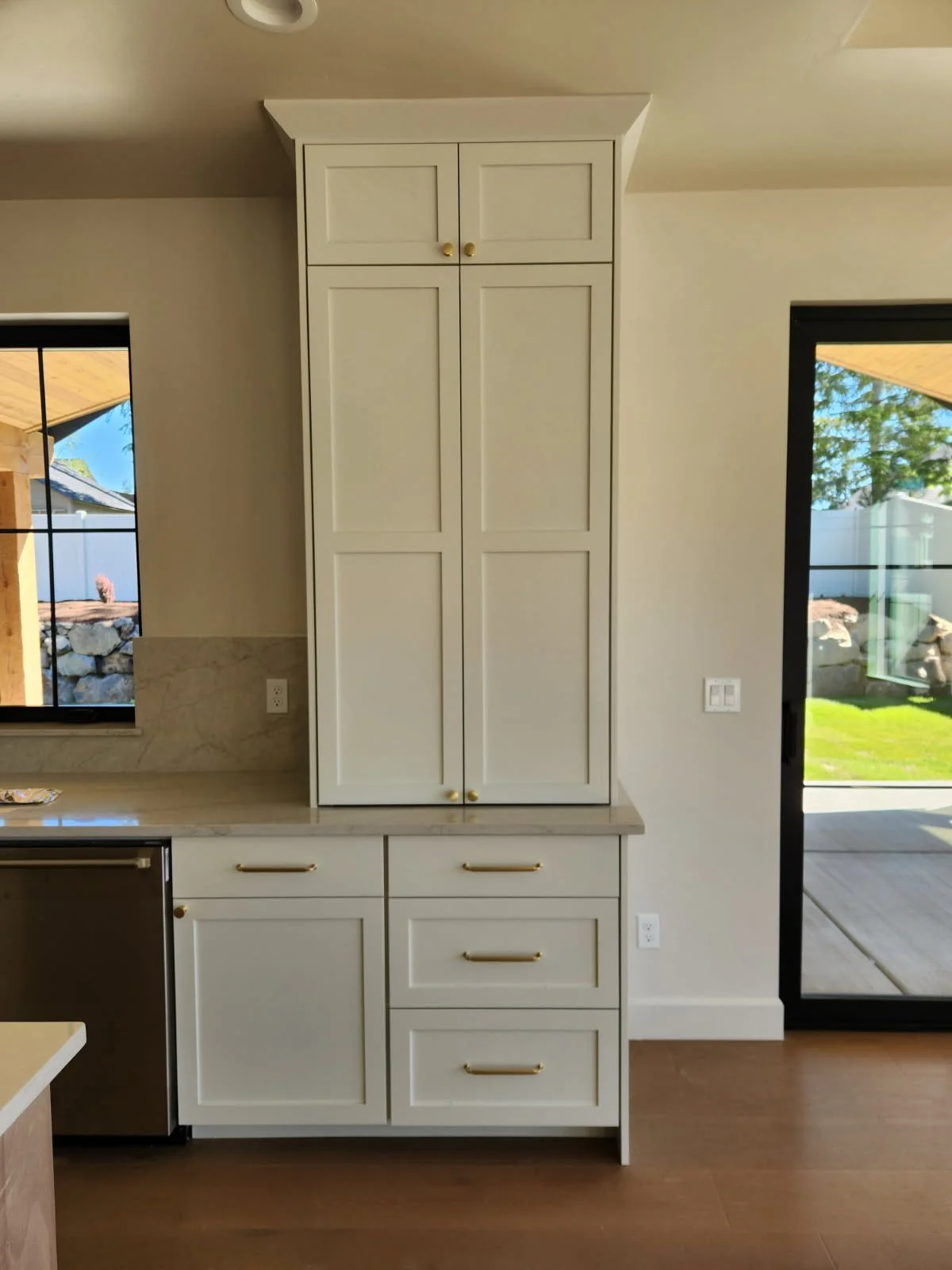 White cabinetry with gold handles in a kitchen, with a window on the left and a sliding glass door on the right, showing a backyard with grass and trees.