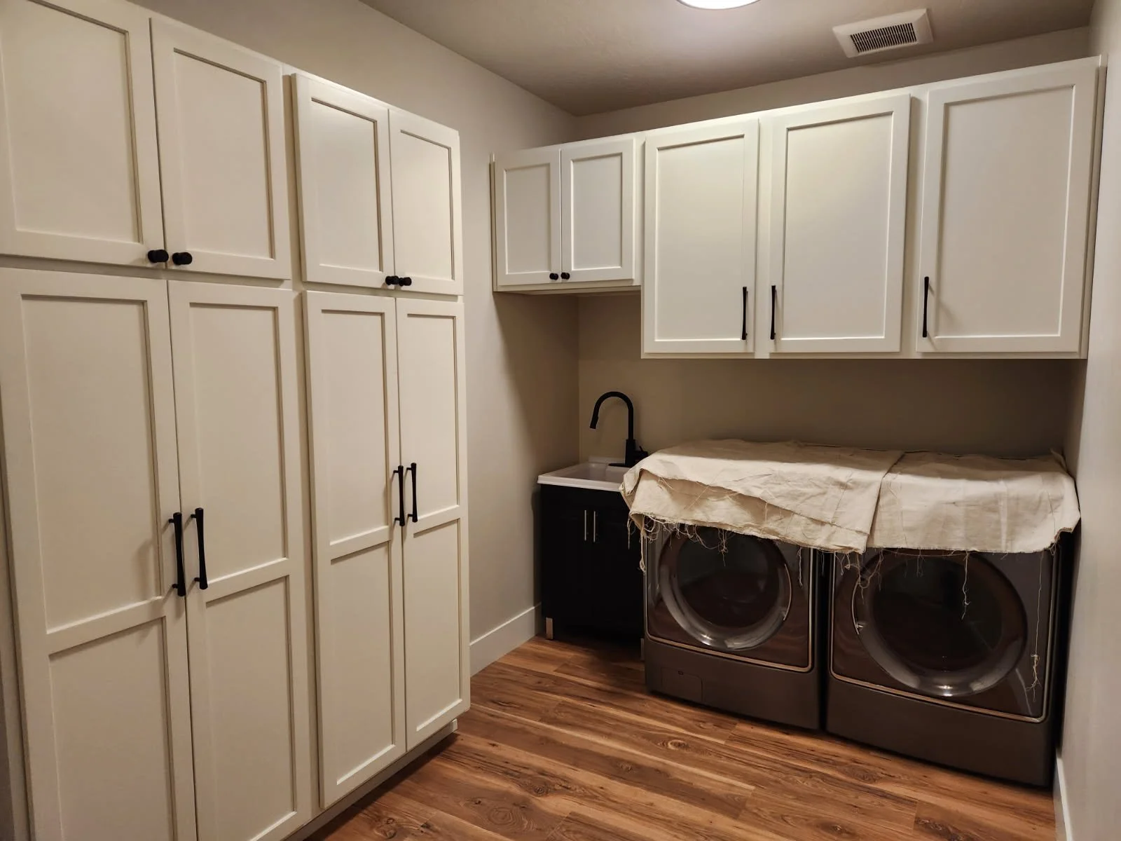 North Idaho Laundry room with white cabinets, a black sink, a washer and dryer covered with a cloth, and a wood flooring.