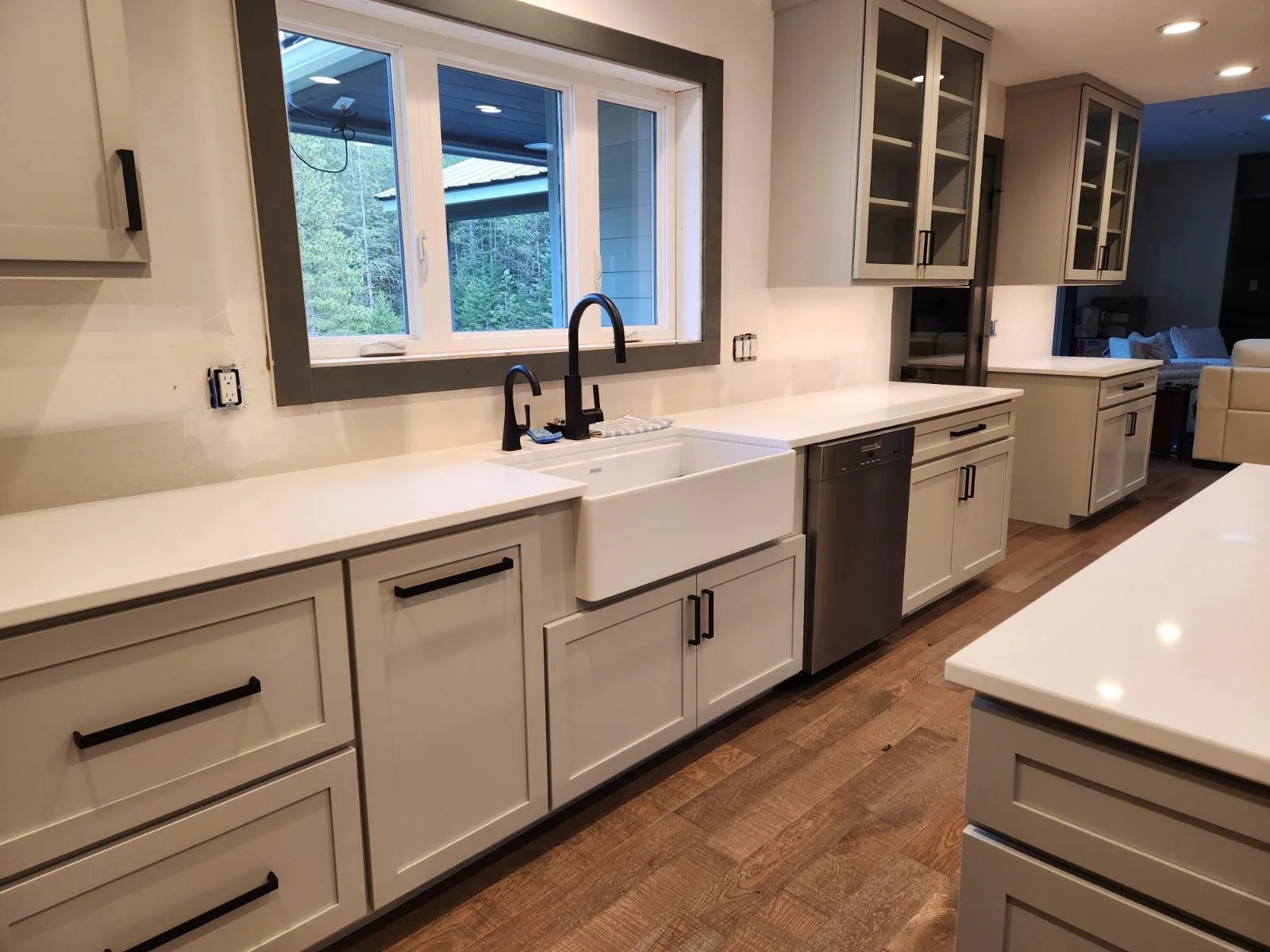 Modern kitchen with white cabinets, black handles, and a large farmhouse sink under a window. The countertop is white, and the kitchen has hardwood floors. There are black faucet fixtures and glass-front upper cabinets. The background shows a living 