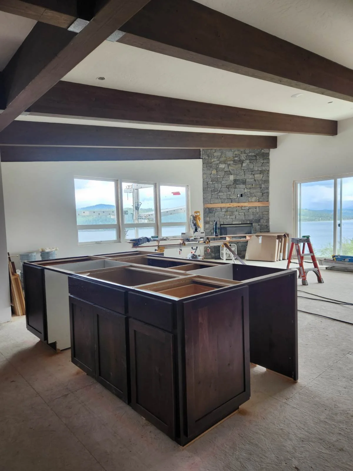 Interior of a house under construction with kitchen cabinets, a stone fireplace, and large windows with a view of water and mountains.