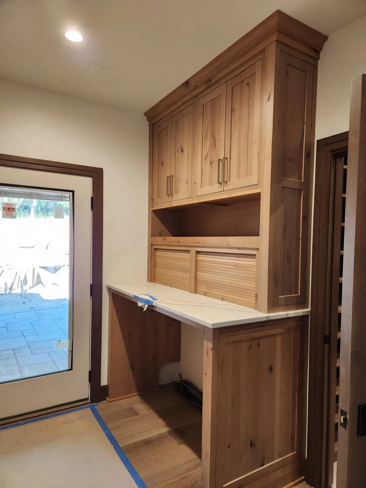 Wooden kitchen cabinets with a countertop and an open space underneath, located near a glass door leading outside, with part of the floor taped off for construction.