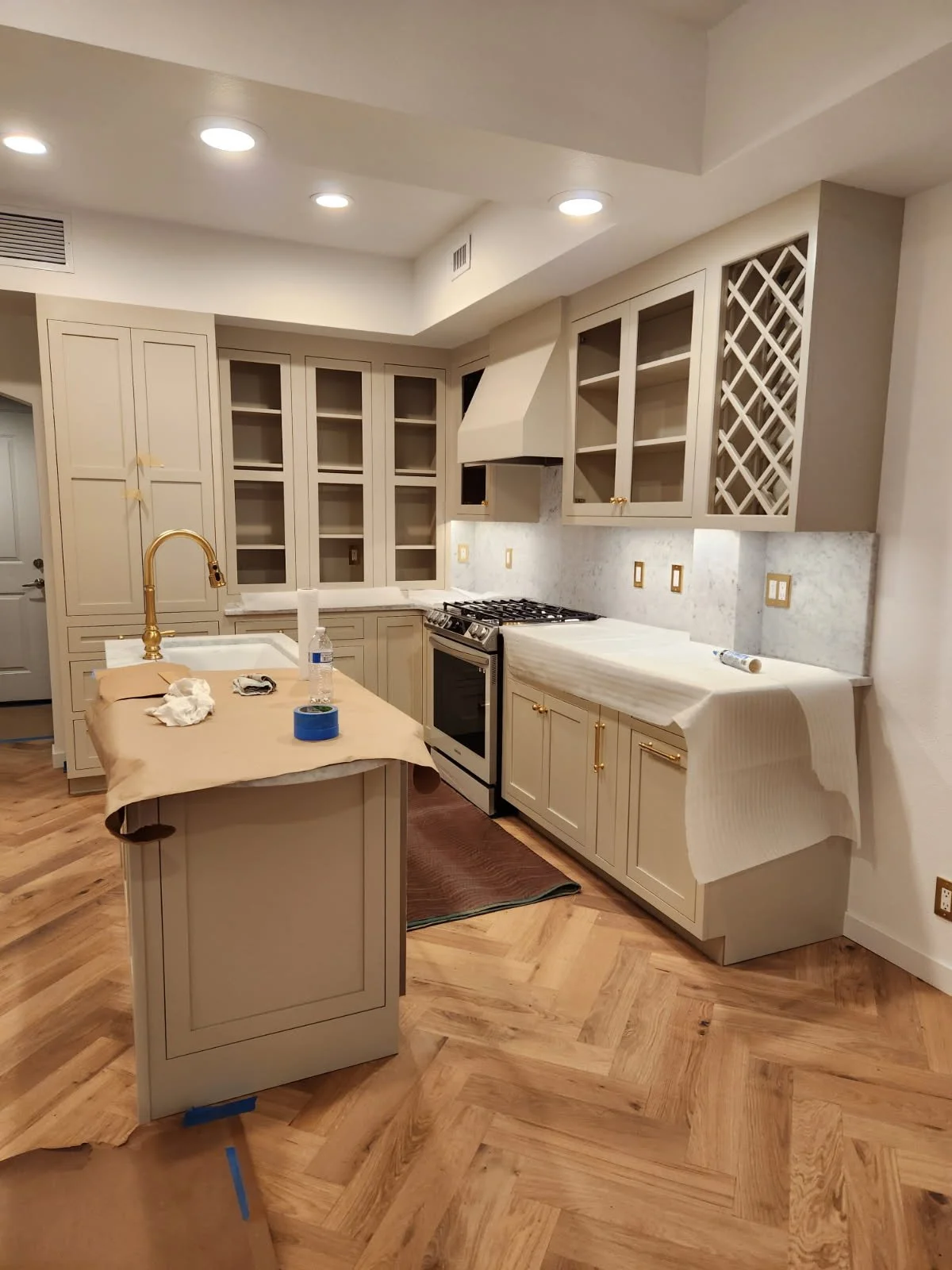 Kitchen under renovation with beige cabinets, marble backsplash, island with a gold faucet, and hardwood floor.