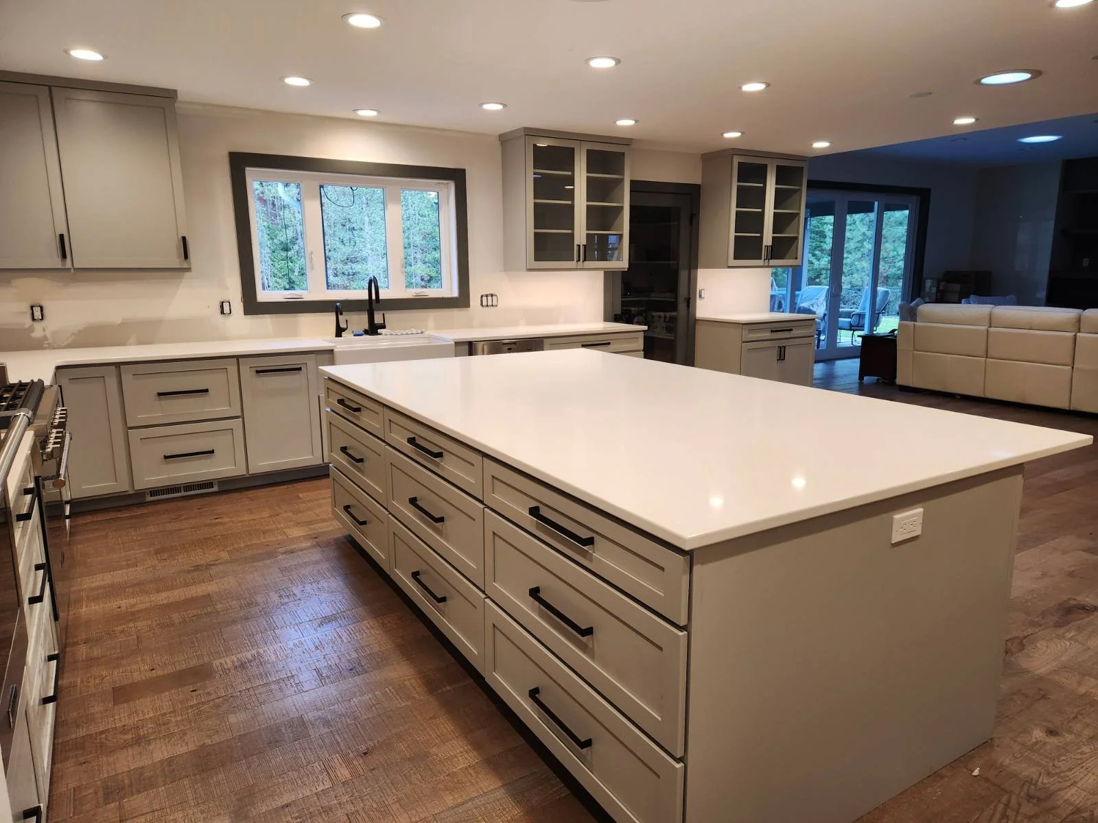 Coeur d'Alene Modern kitchen with a large island, white countertops, gray cabinets, hardwood flooring, and a window above the sink with a view of trees outside.