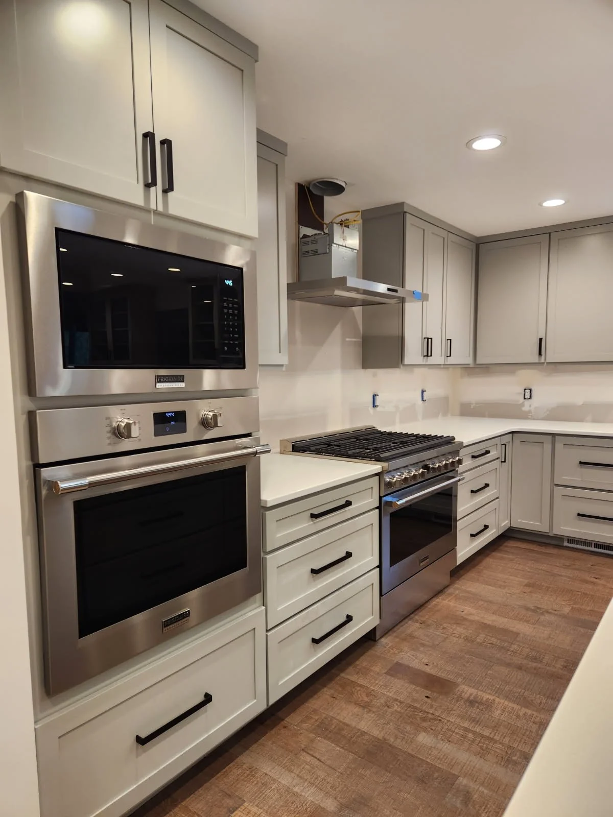 Kitchen with white cabinets, stainless steel oven and microwave, stove with oven, hardwood floor, and unfinished wall area.
