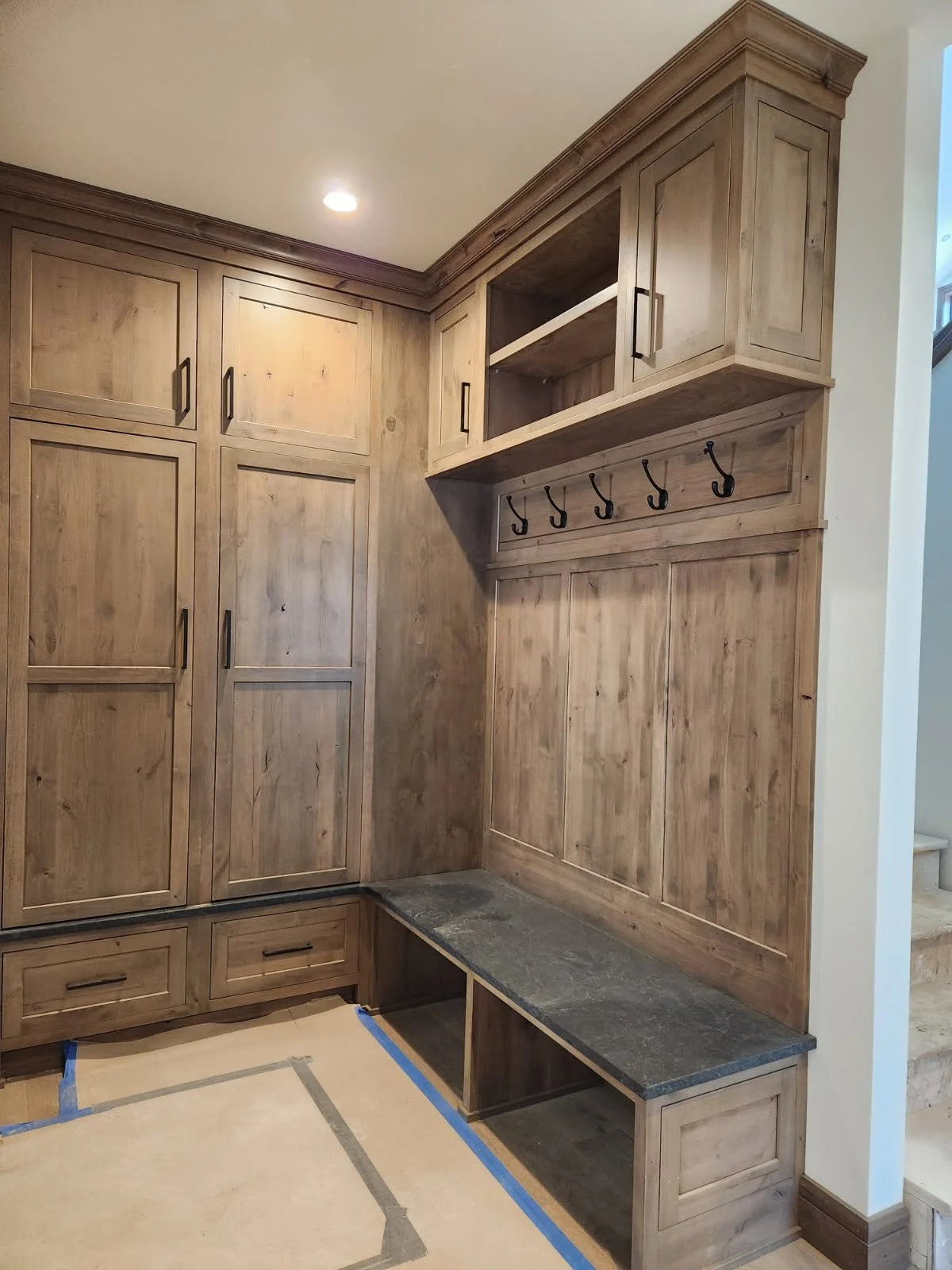 View of a built-in wooden mudroom storage unit with cabinets, hooks, and a bench with a stone top, adjacent to a staircase.