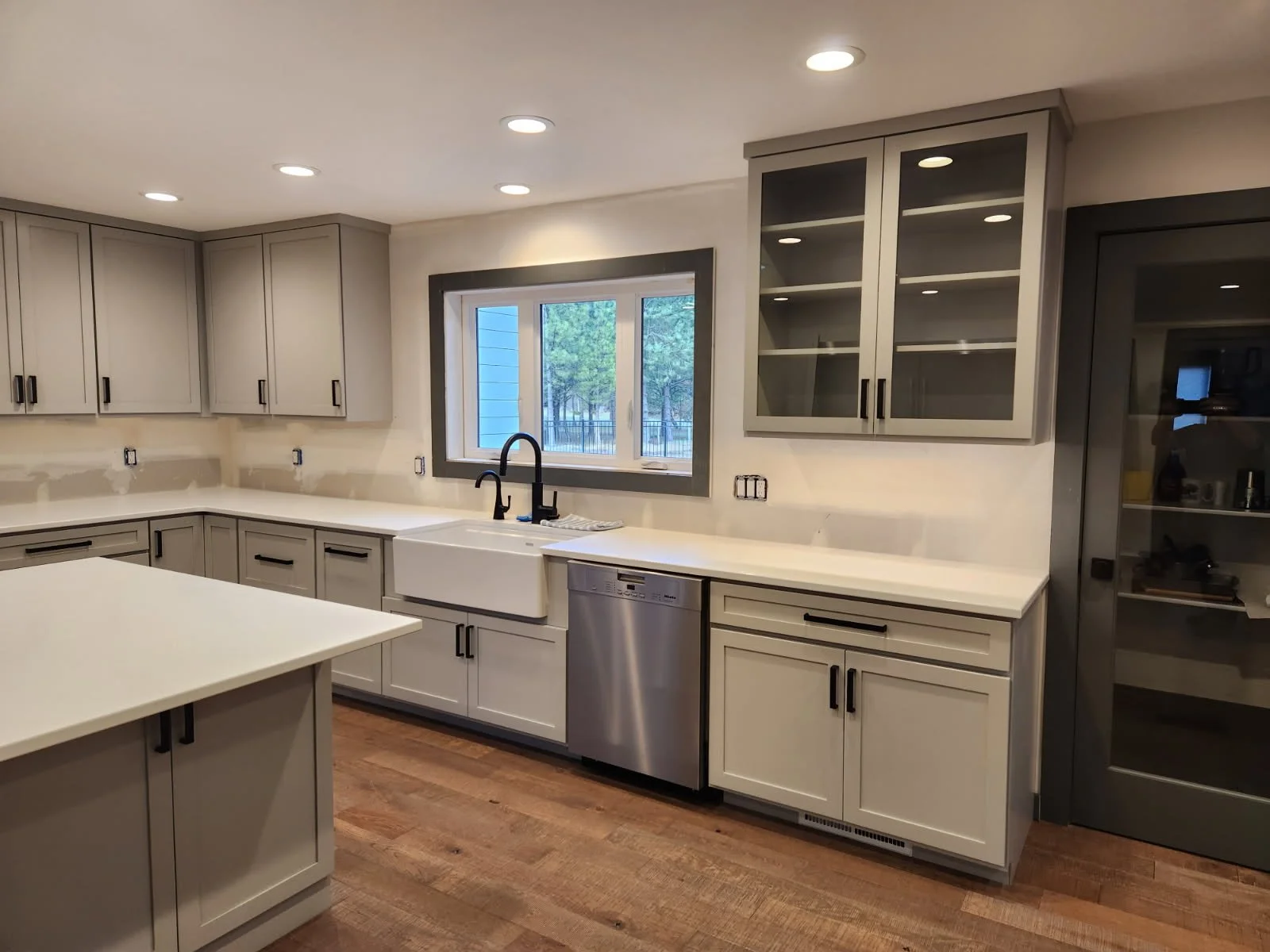 A clean, modern kitchen with white countertops, gray cabinets, a white farmhouse sink, a stainless steel dishwasher, and a window overlooking trees. There are cabinets and drawers, with some opened shelves, and a wooden floor.