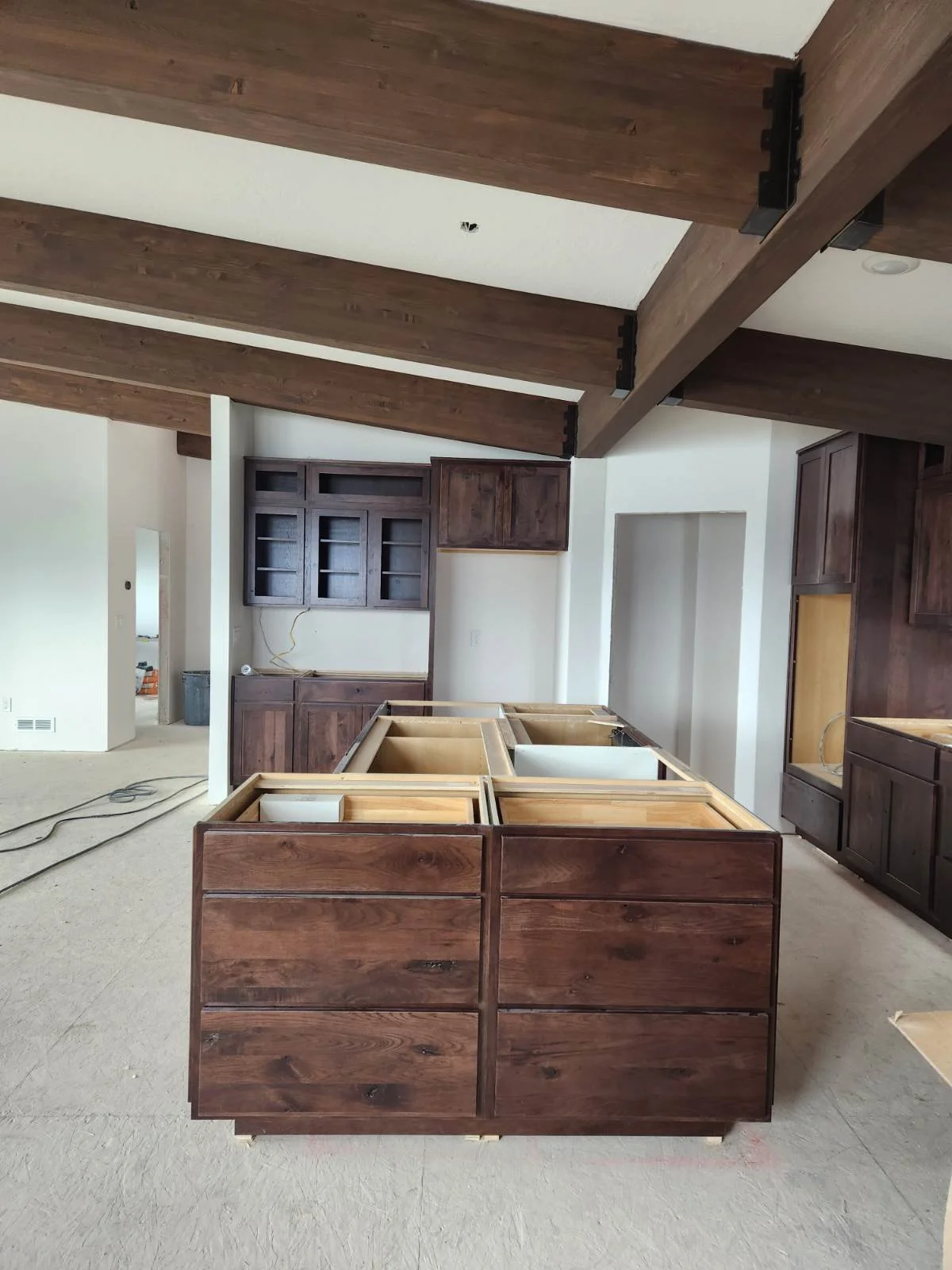 Kitchen under construction with dark wooden cabinets and an island, open shelves on the wall, and exposed ceiling beams.