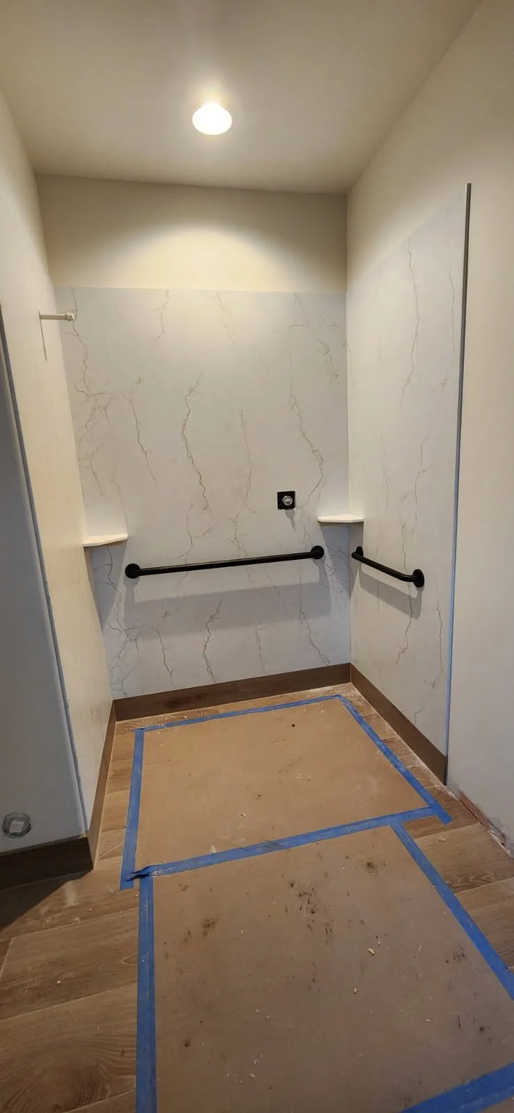 Empty shower stall with marble-patterned walls, two black safety grab bars, small white corner shelves, a ceiling light, and taped-off floor indicating ongoing renovation.