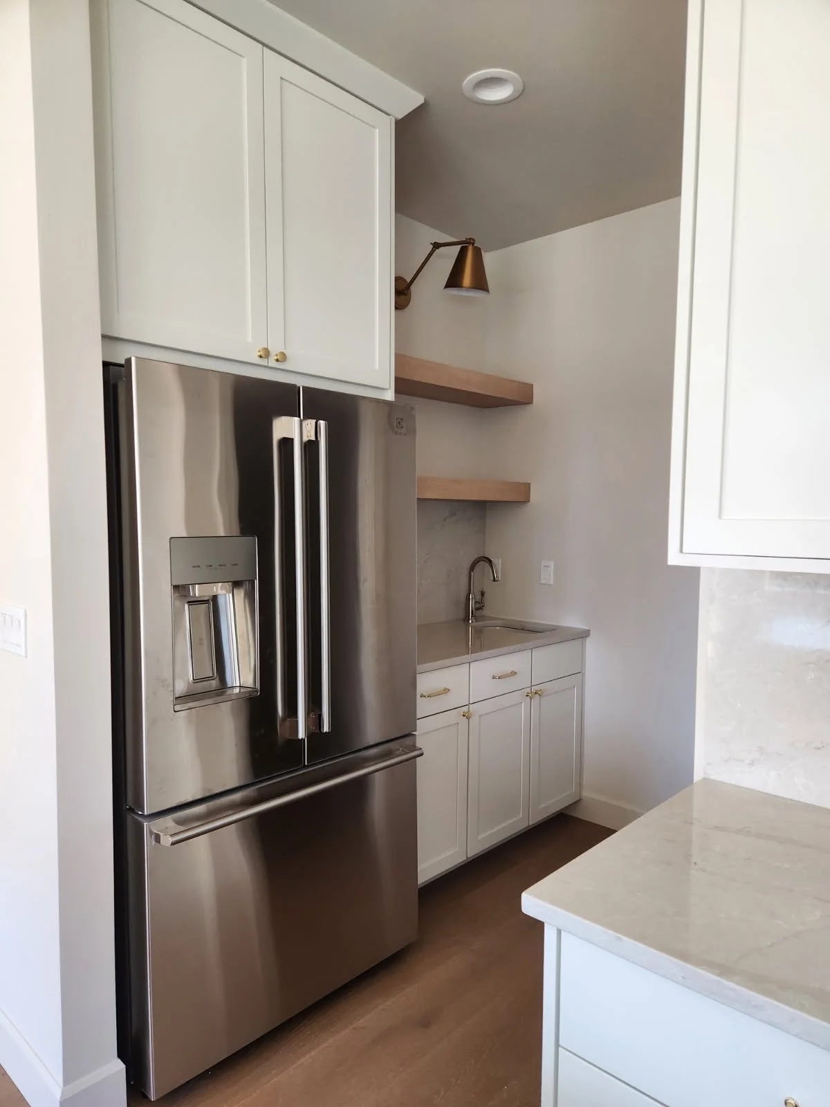 Kitchen with a stainless steel refrigerator, white cabinets, wooden floating shelves, a small sink, and gold hardware.