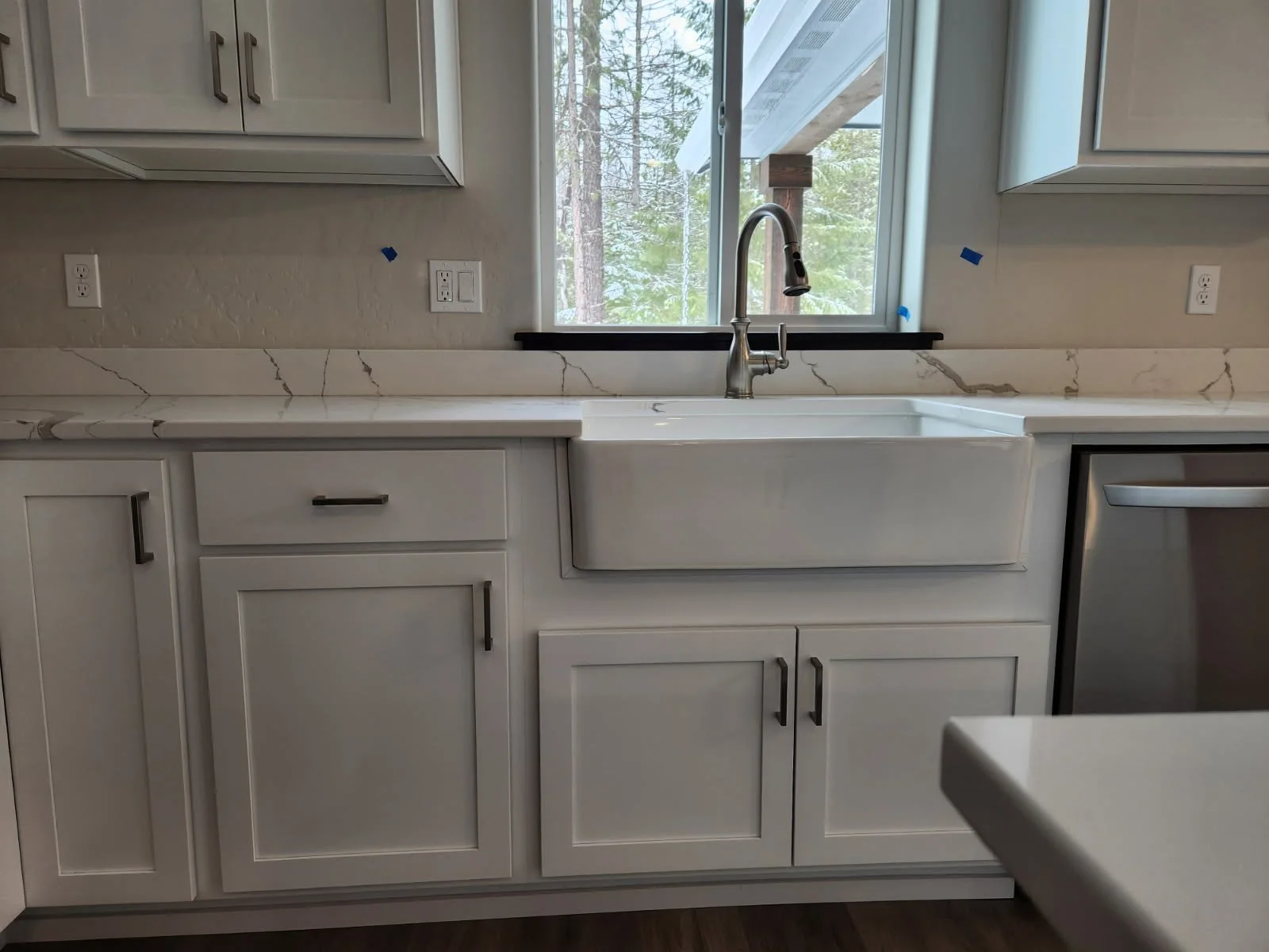 Coeur d'Alene Modern white kitchen with a farmhouse sink, marble countertop, and a window above the sink showing trees outside.