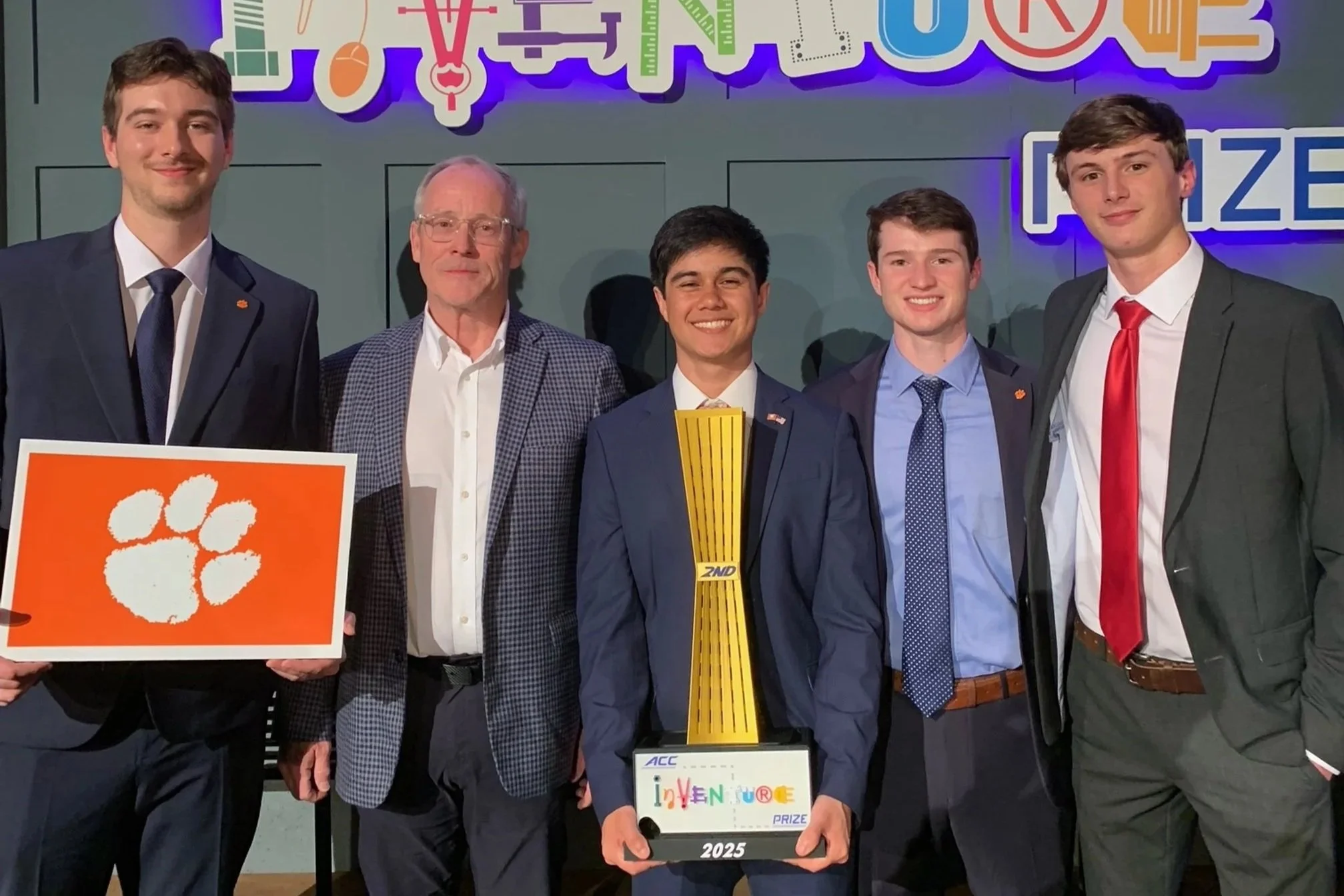 Group of five young men and one older man in suits, standing together at an awards ceremony. One young man in the center holds a trophy with colorful text that reads 'inventure' and the year 2025. The background has a colorful sign with various scientific and technological icons and the word 'PRIZE.' One person on the far left holds a placard with a white paw print on an orange background.