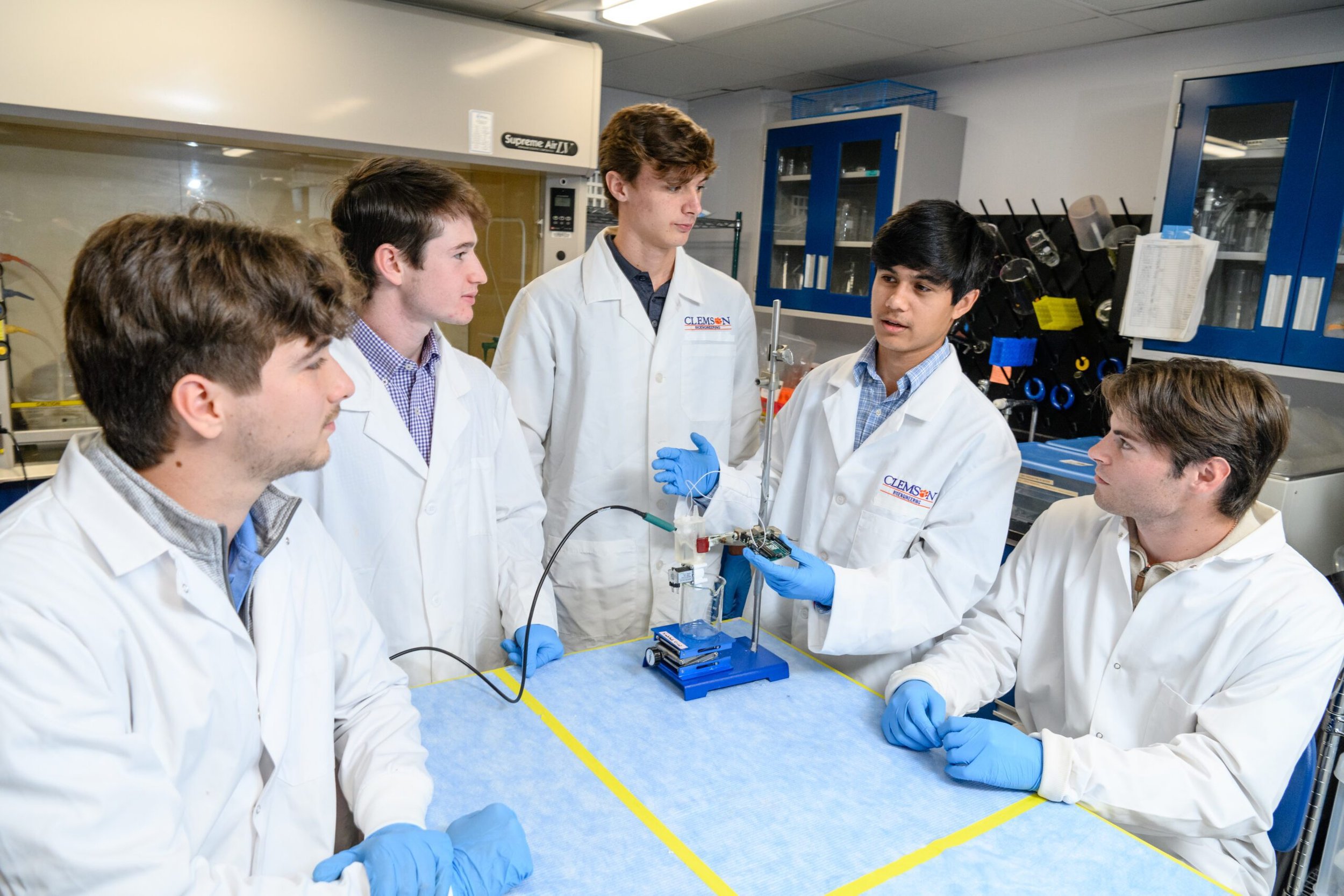 Group of six young scientists in lab coats and blue gloves gathered around a scientific experiment in a laboratory. One of them is explaining the apparatus to the others.