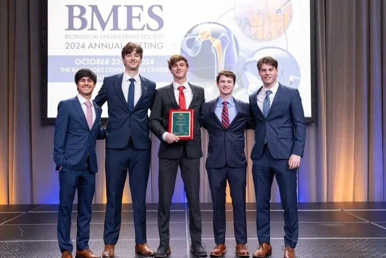 Five young men in suits standing on stage at a conference, with the banner behind them reading 'BMES 2024 Annual Meeting, October 23-25, 2024, The Baltimore Convention Center.' The man in the center holds a plaque, indicating an award or recognition.