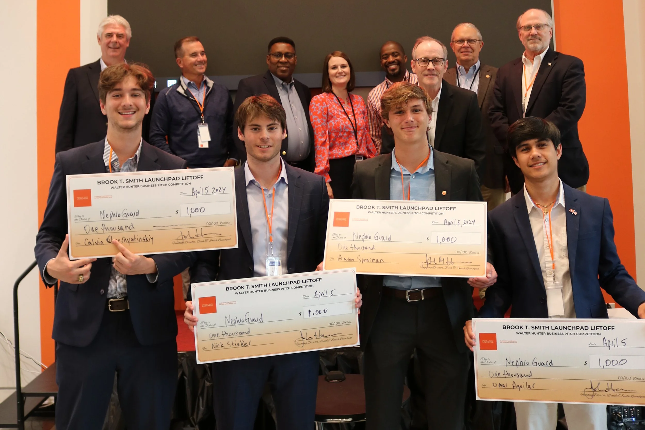 Group of young men and older men standing on stage, holding large checks with the date April 15, 2024, at the Walter Hunter Business Pitch Competition, celebrating their awards.