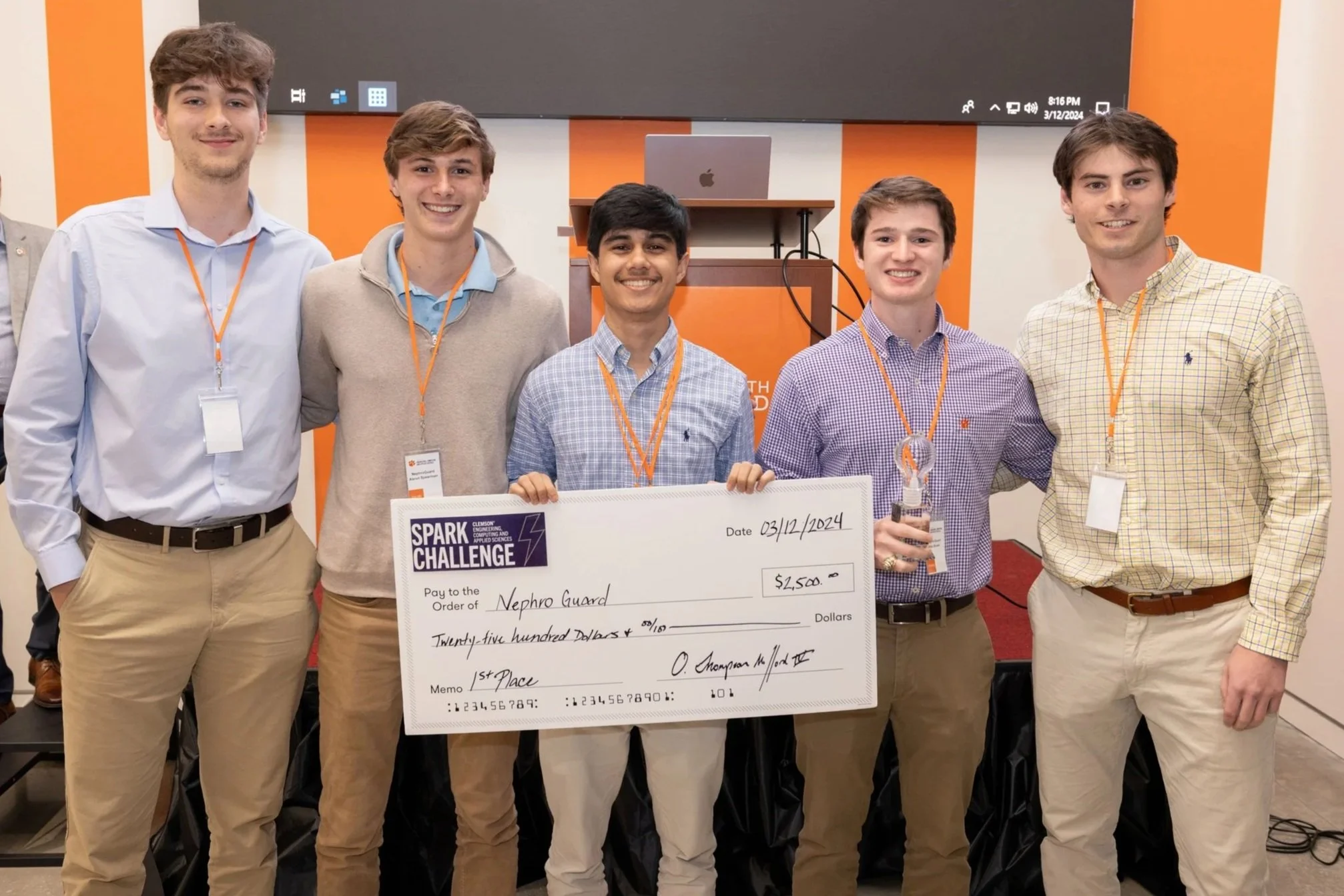 Six young men standing together, holding a large check and a trophy, at an award ceremony or competition event.