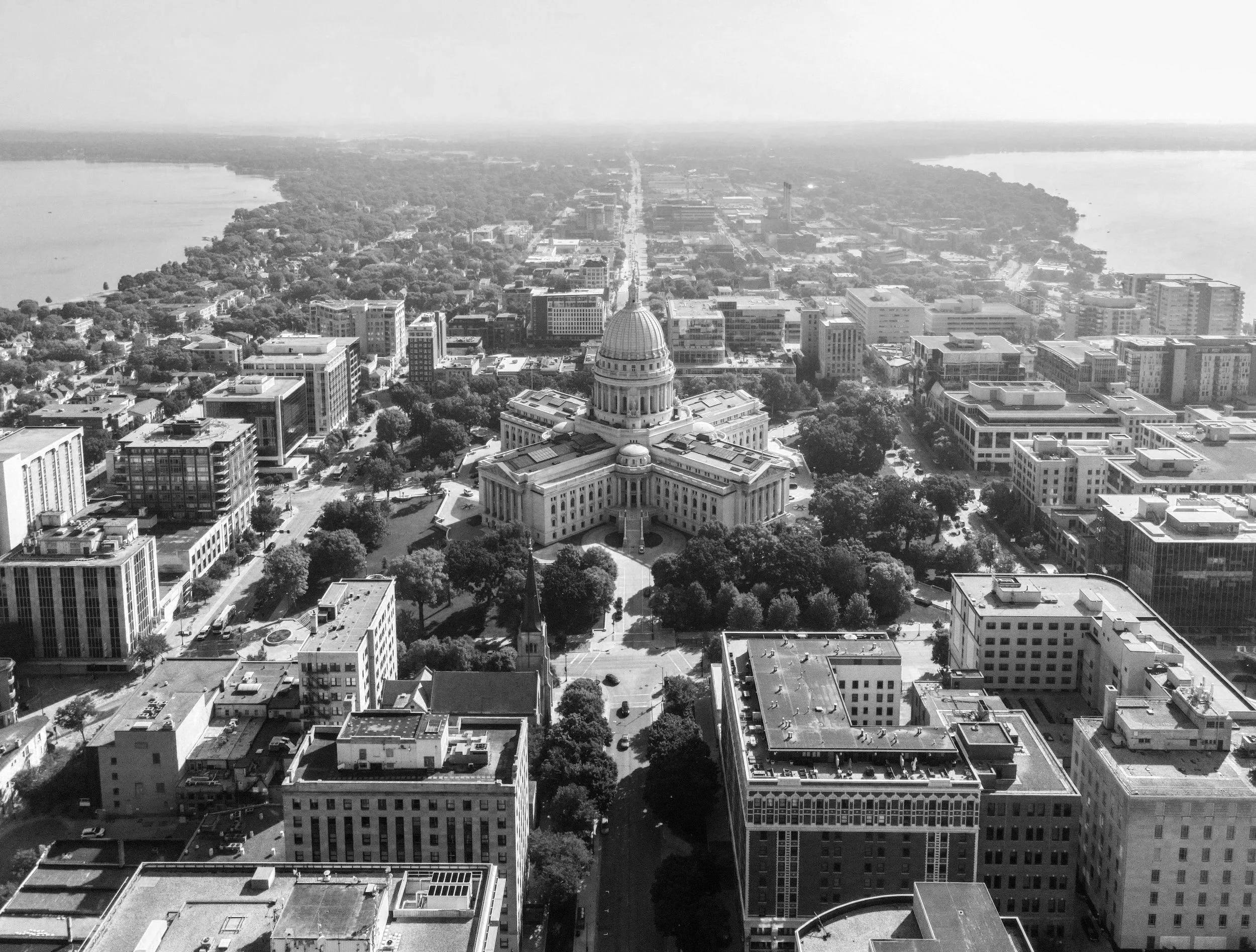 A black-and-white aerial view of a cityscape featuring a prominent domed government building in the foreground, surrounded by modern high-rise buildings, with a river on the left and a lengthy straight road extending into the distance.