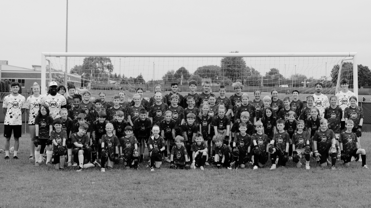 Group photo of children and coaches on a soccer field with goals and trees in the background, all wearing sports uniforms.