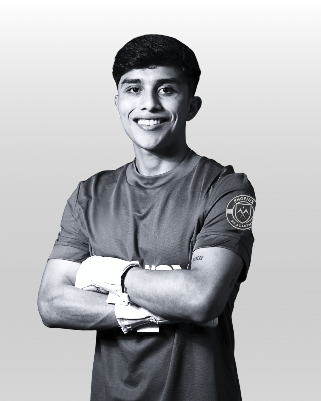 Young man with military-style haircut smiling, wearing a sports jersey with a patch on the sleeve that reads 'Phoenix GK Academy', crossing his arms, standing against a plain background.
