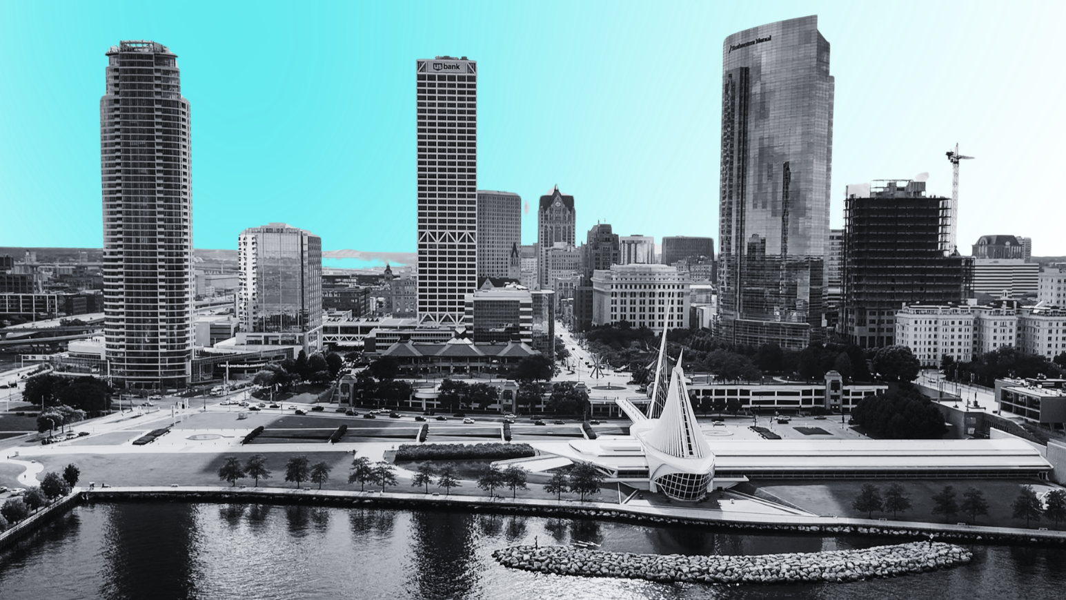 Aerial view of a waterfront cityscape with modern skyscrapers, a unique boat-shaped building, and a body of water in the foreground.