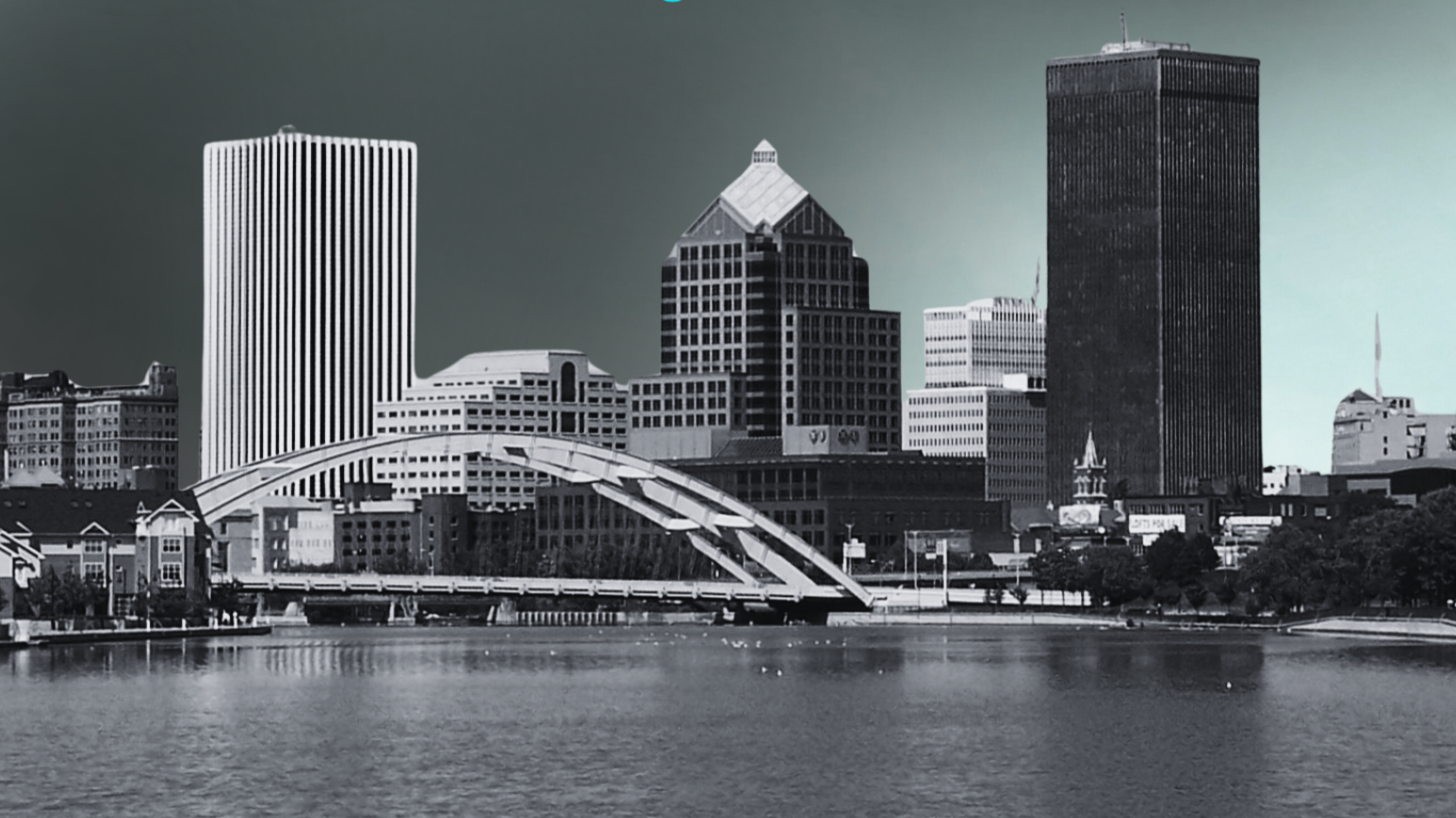 Black and white photo of a city skyline with tall modern buildings, a bridge over water in the foreground, and a clear sky.