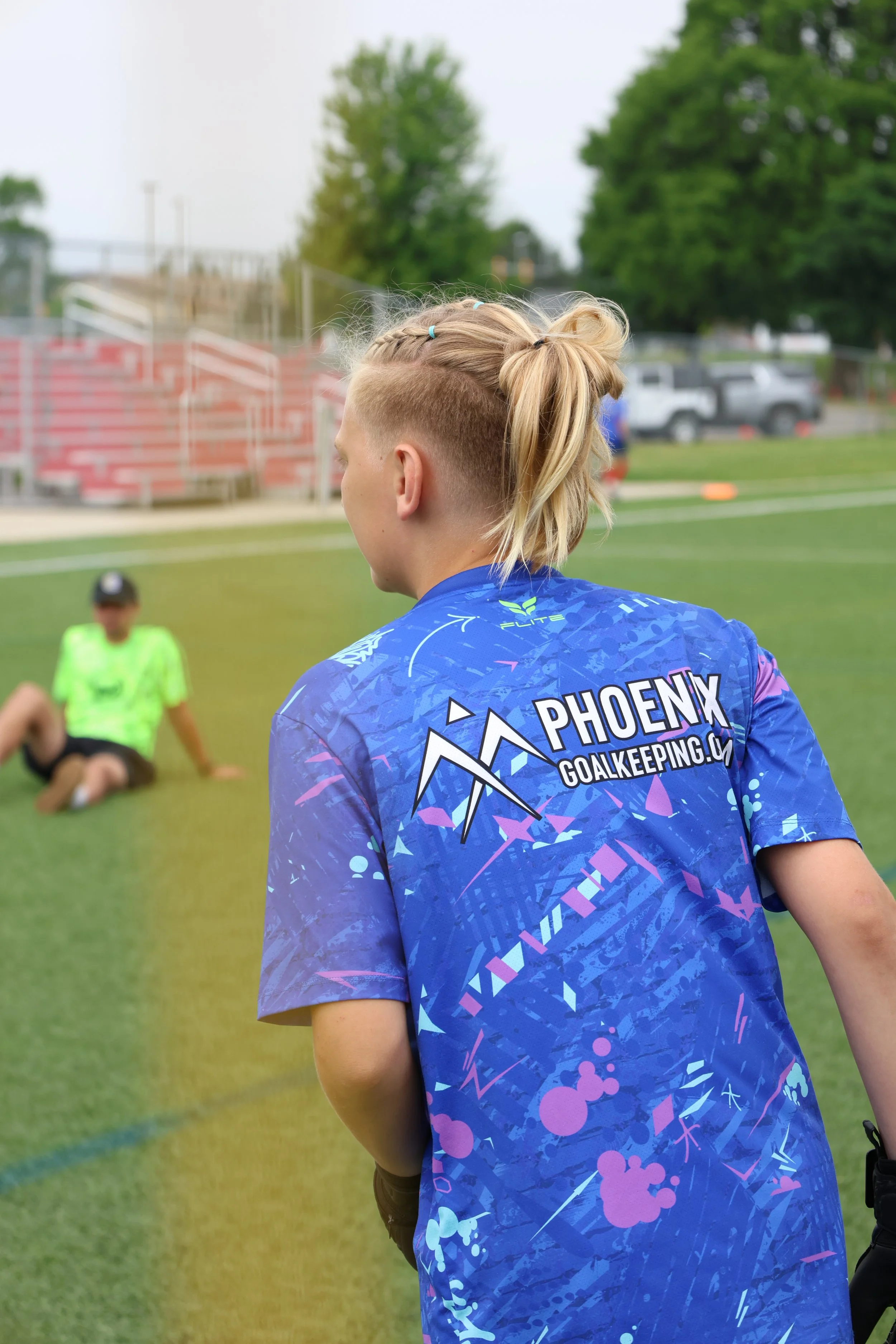 A male soccer player with a partly shaved hairstyle and tied-up blonde hair is standing on a soccer field, wearing a blue team jersey with the words "PHOENIX GOALKEEPING.COM" printed on the back.
