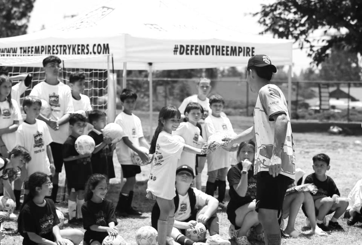 Children in sports uniforms receiving an award or trophy from an adult during a youth soccer event under a tent with banner and hashtag.
