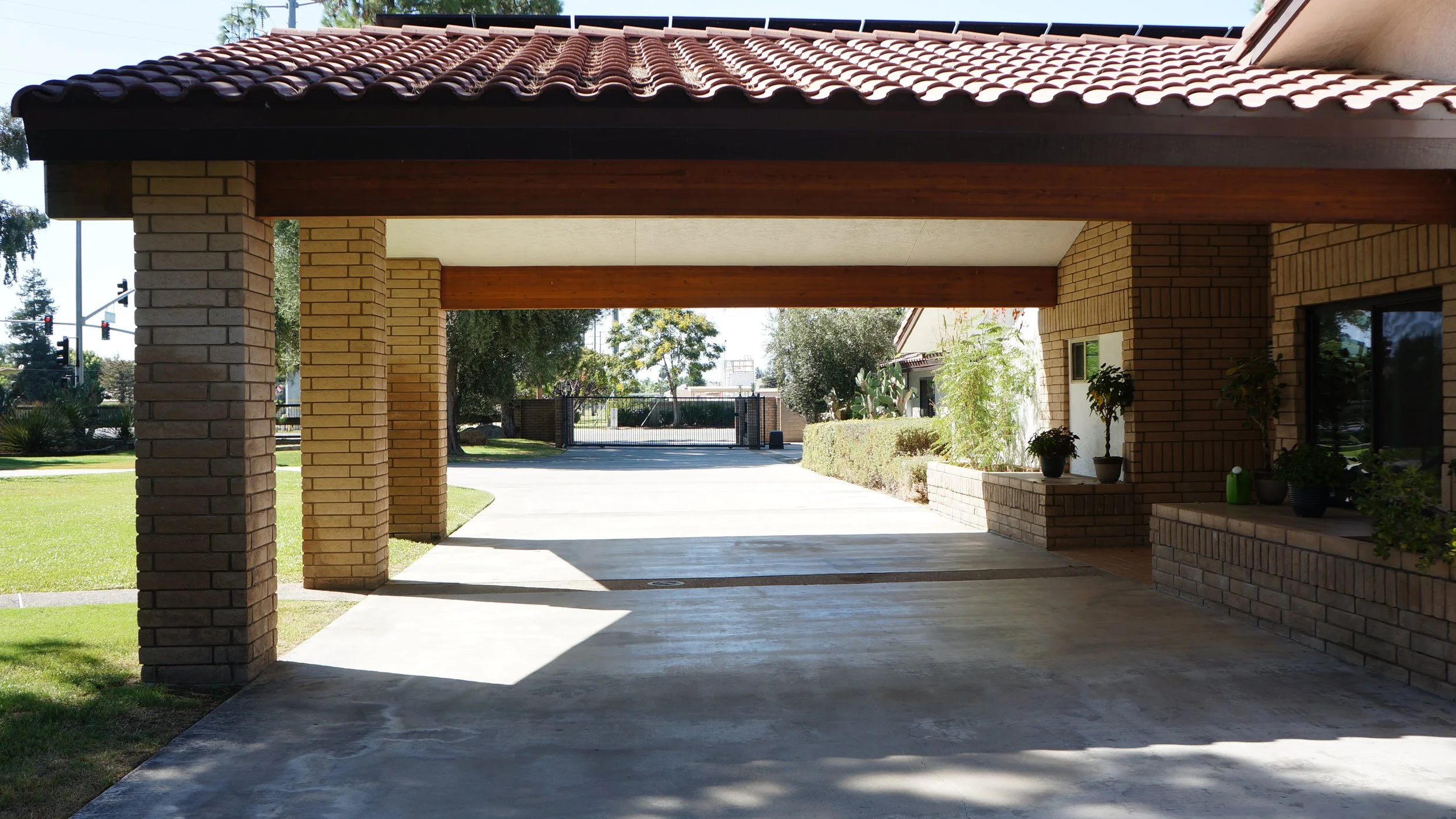 A covered parking area with brick pillars, a tiled roof, and a concrete floor, overlooking a green lawn, trees, and a gate in the background.