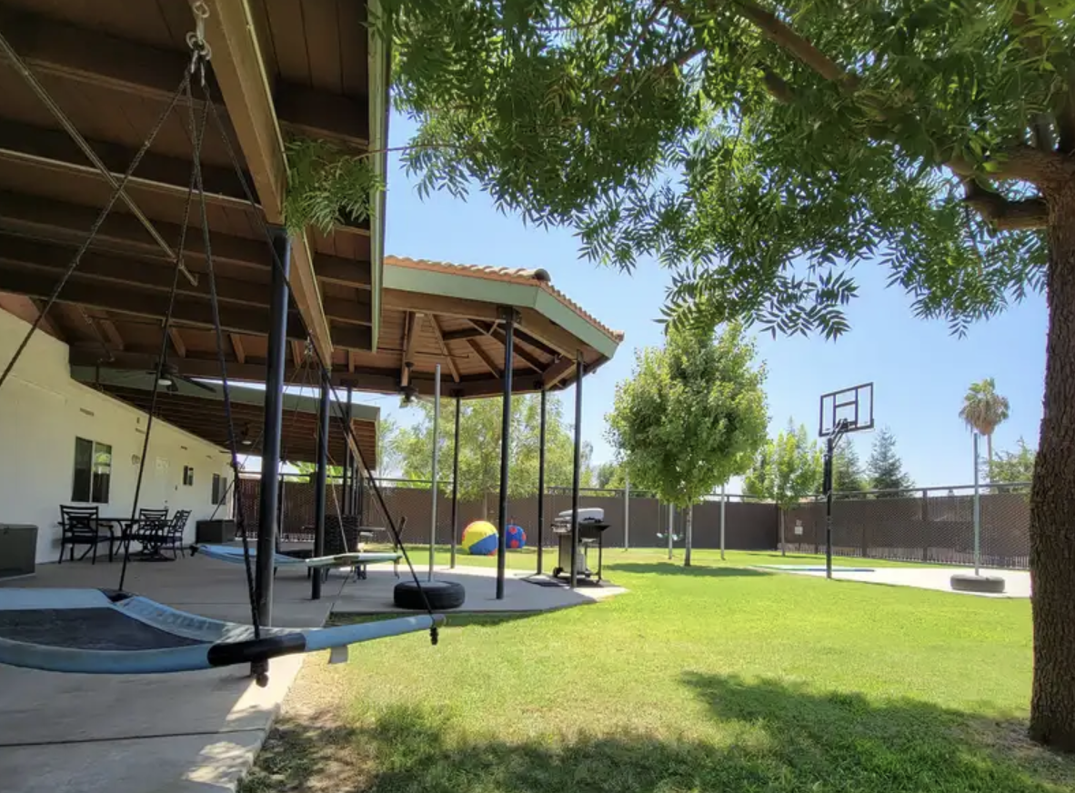 Residential backyard with swing set, patio, table and chairs, barbecue grill, trees, and a basketball hoop on a sunny day.