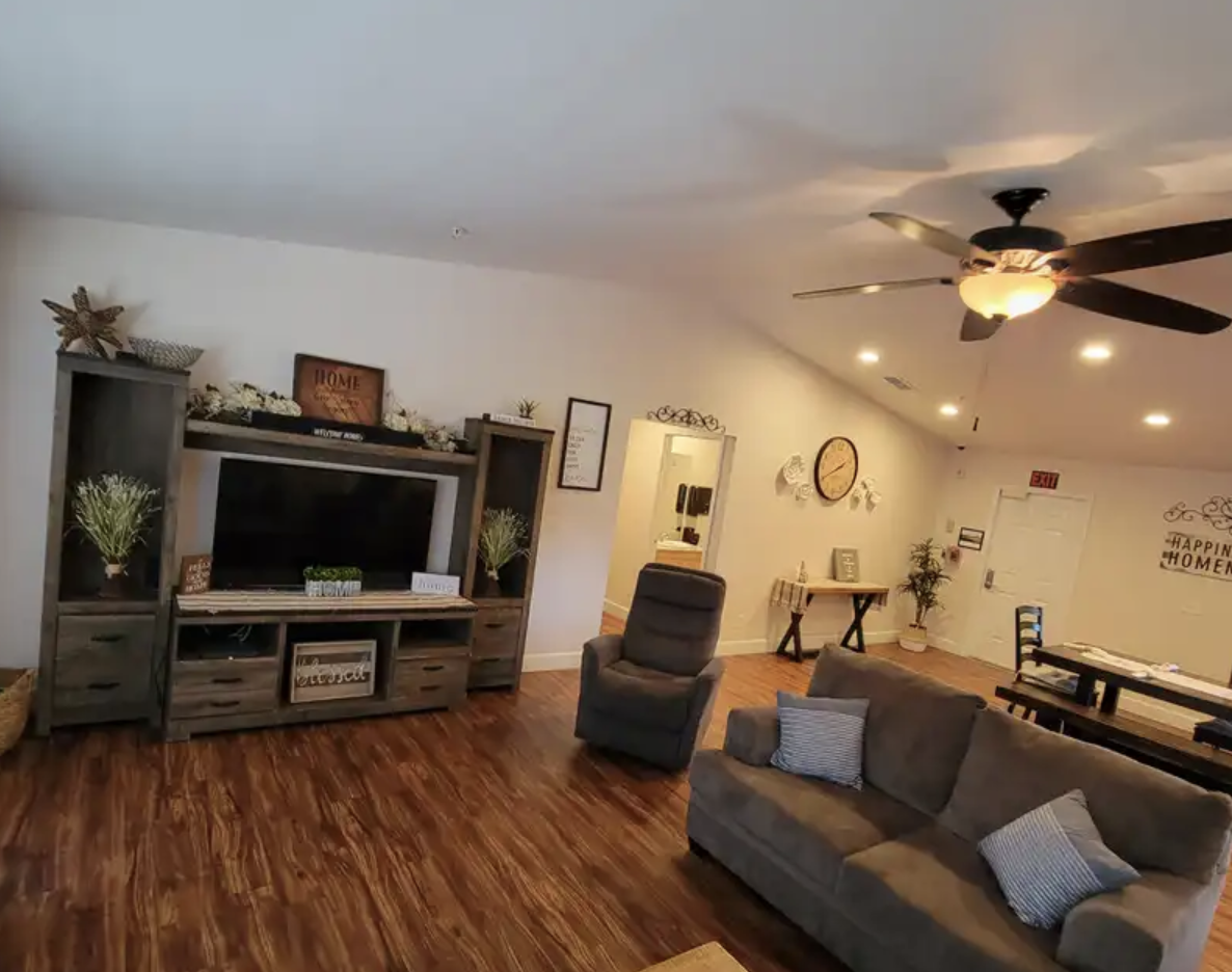 Living room with gray sofa, recliner, wall clock, television on gray entertainment center, ceiling fan, wood floor, and wall decorations.