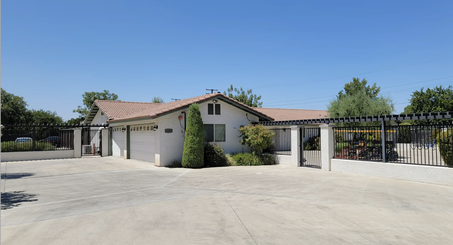 A single-story house with a red tile roof, two-car garage, white exterior walls, surrounded by a metal fence, with some trees and landscaping in front and a spacious concrete driveway.