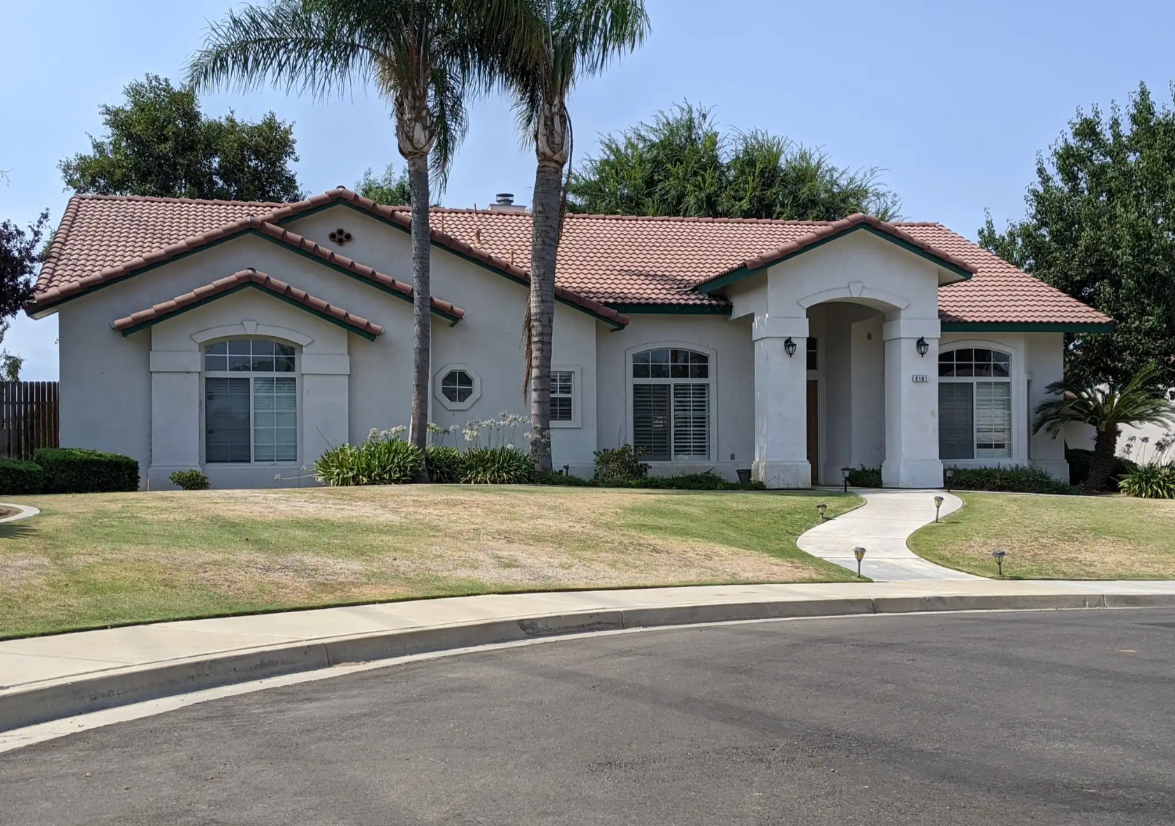 Front view of a white house with a red-tiled roof, large windows with white shutters, a curved pathway leading to the front door, palm trees, and landscaped lawn.