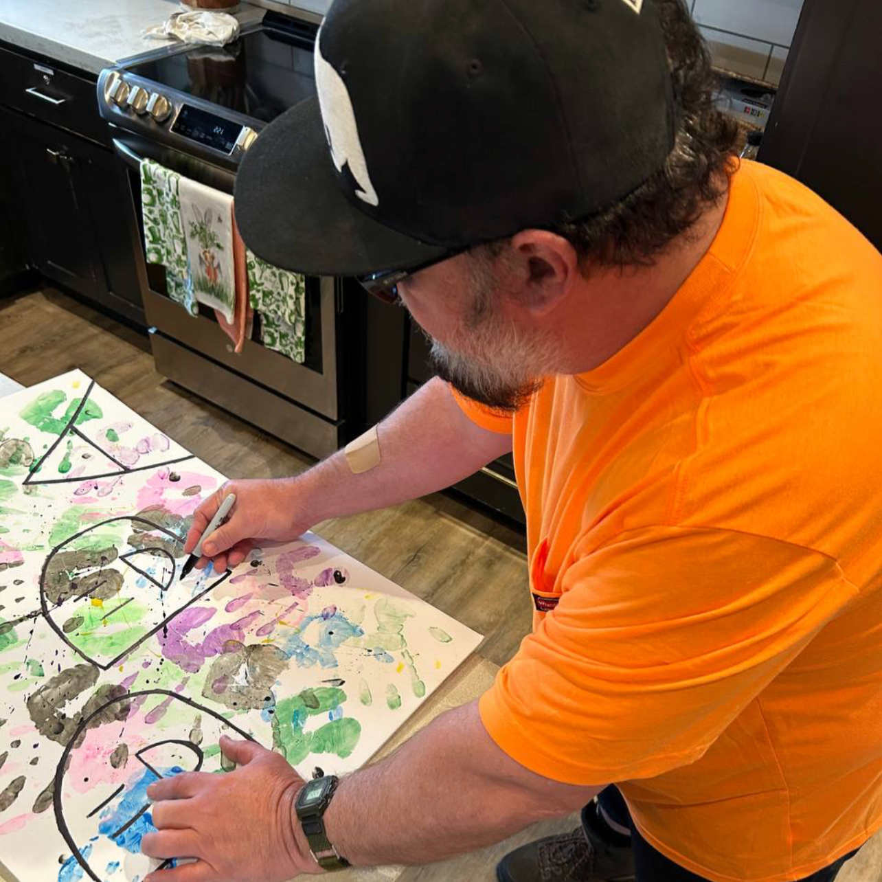A man with a beard and glasses, wearing a black baseball cap and an orange shirt, is drawing on a large sheet of paper with colorful paint splatters and black outlines, in a kitchen with black cabinets and a stove.