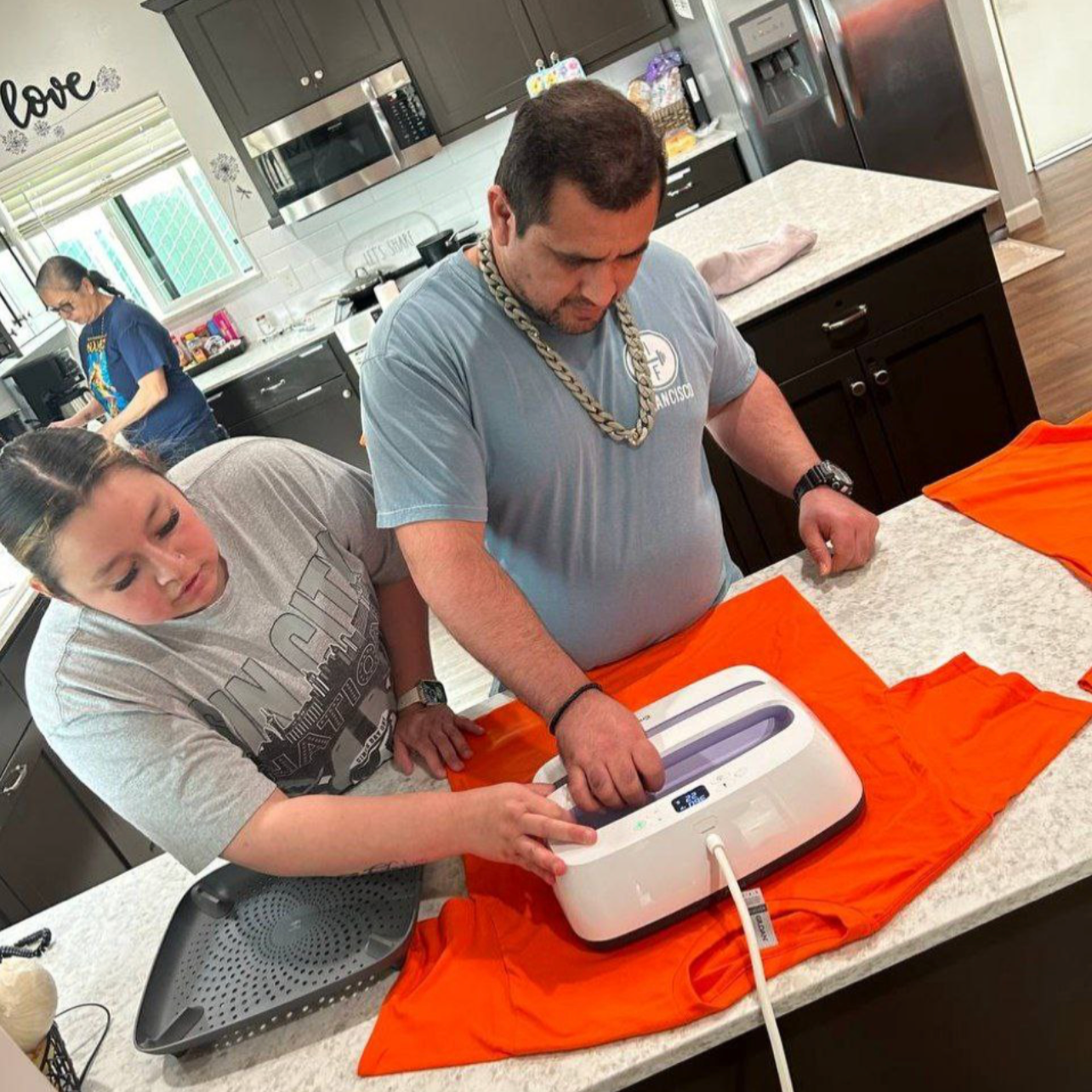 Two people in grey tops are pictured using an iron to apply appliques to a bright orange tee shirt. The person on the right is wearing a shiny gold chain and a black watch.