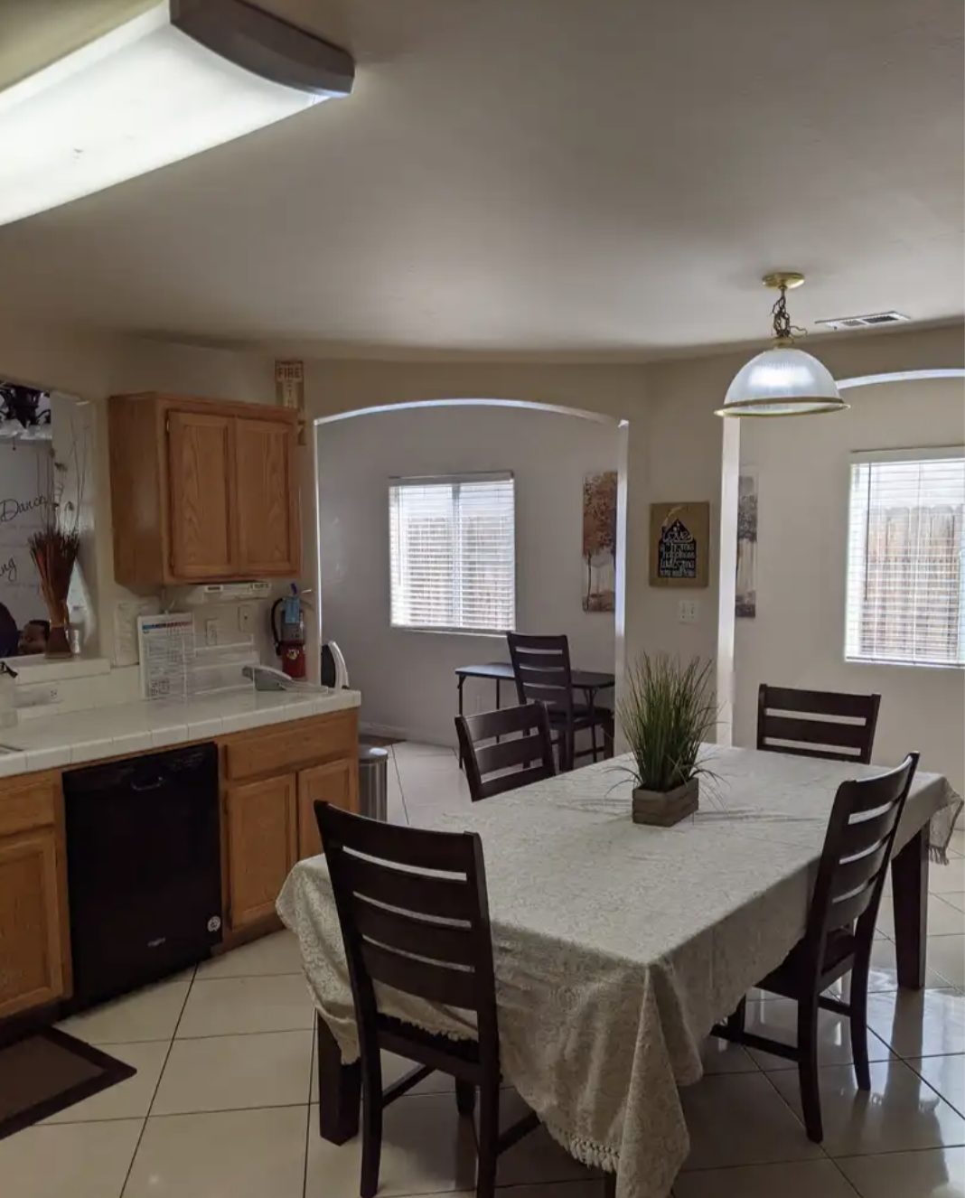 Dining area with a table covered in a lace tablecloth, surrounded by six dark wooden chairs, with a small potted plant in the center, next to a kitchen with wooden cabinets, a window with blinds, and additional windows in the background.