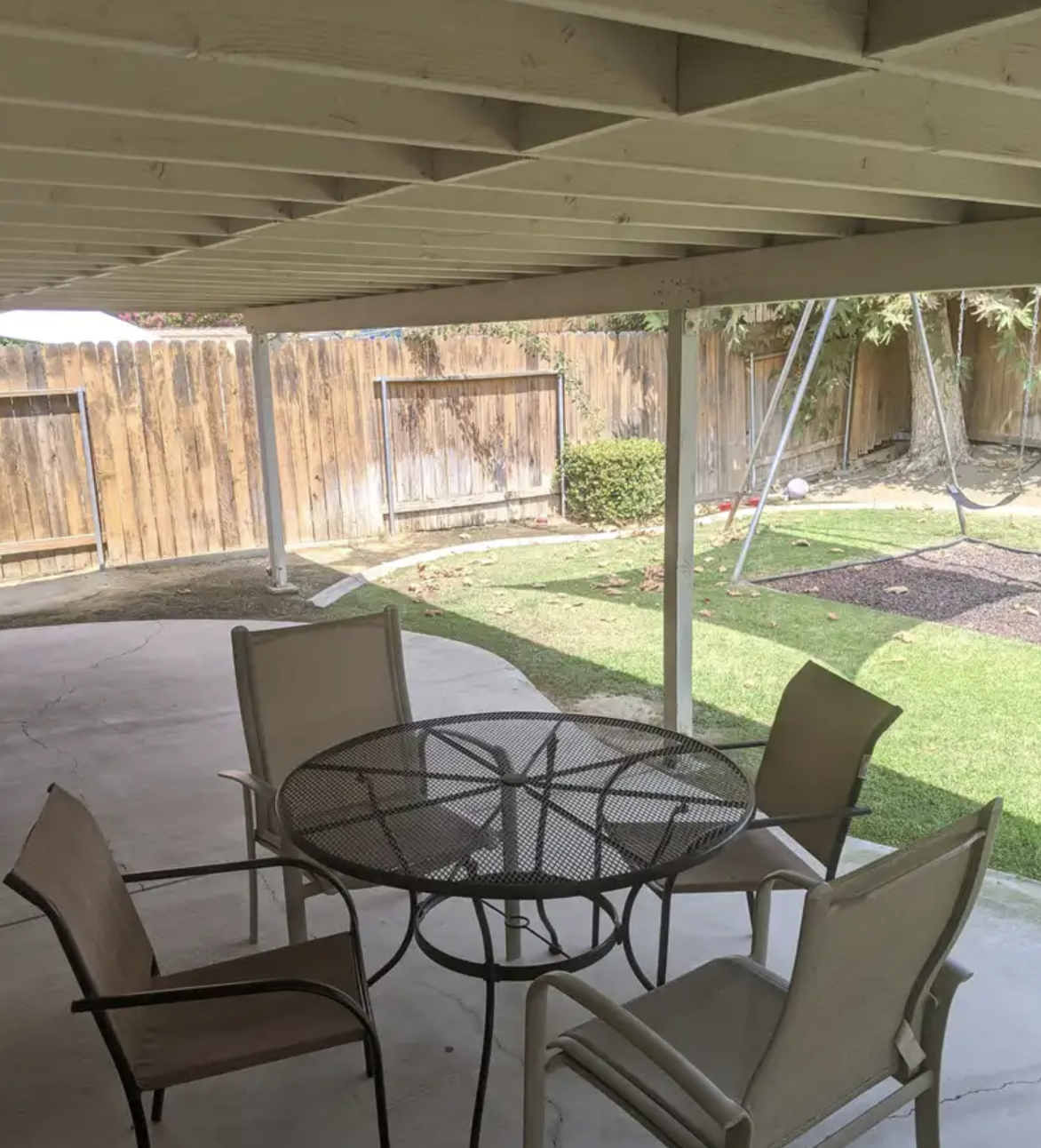 A backyard patio area with a round metal table and four beige chairs underneath a wooden porch cover. The yard has green grass, a wooden fence, a small bush, and a swing set with a black seat on the right side.