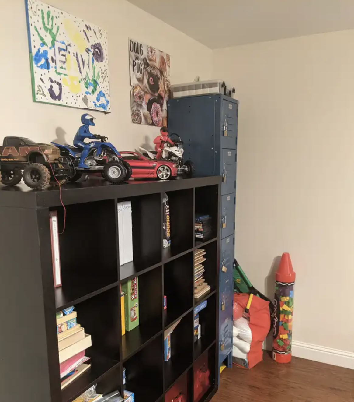 A black bookshelf with multiple compartments topped with toy vehicles, including a blue motorcycle, a pink sports car, a white motorcycle, and a black off-road vehicle. Two posters are on the wall above, one with colorful handprints and the name 'Eli
