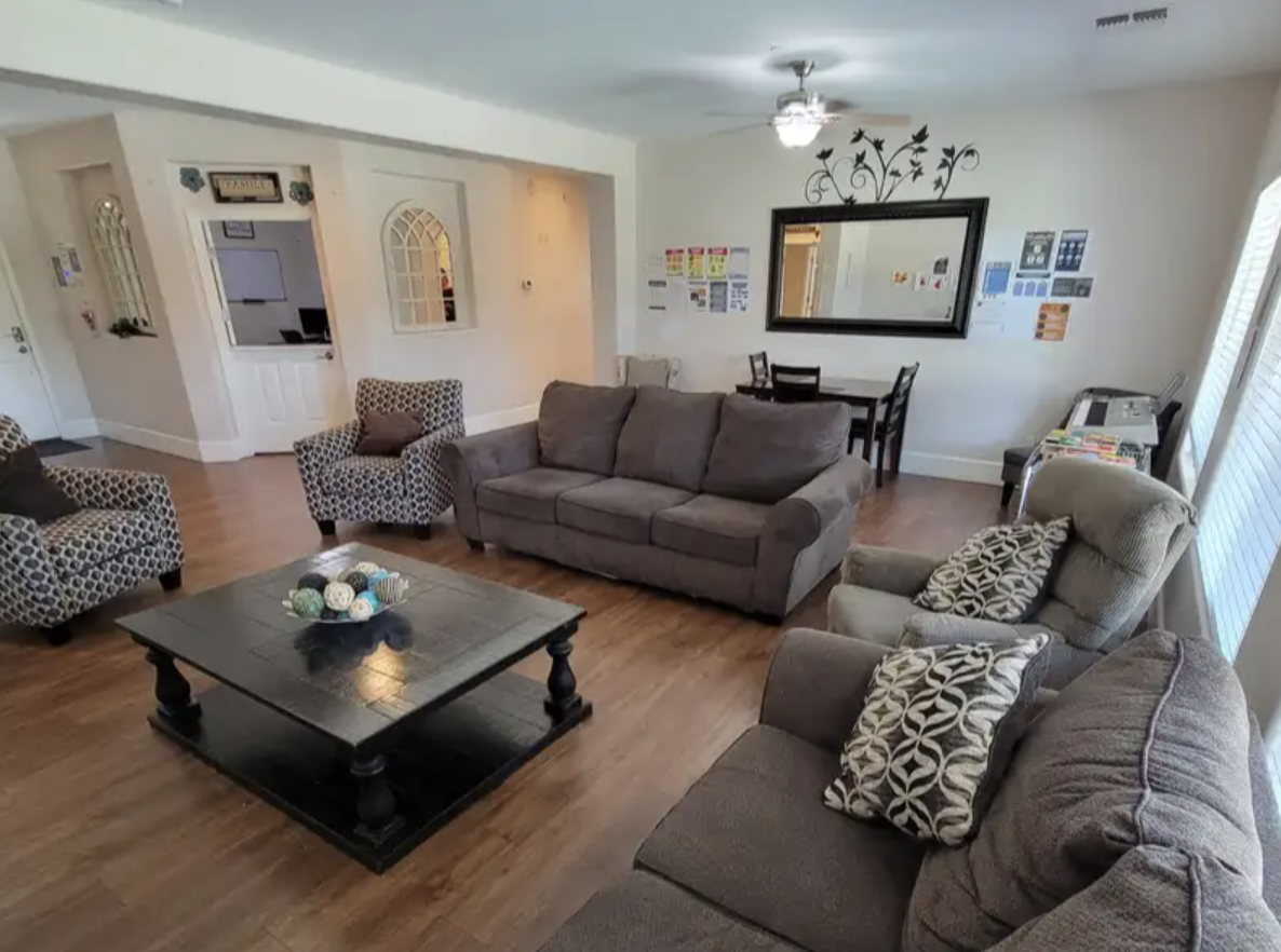 Living room with a gray sofa, armchairs, a black wooden coffee table with decorative yarn balls, a dining table with four chairs, and wall decorations including a large mirror and framed art. Wooden flooring and a ceiling fan are also visible.