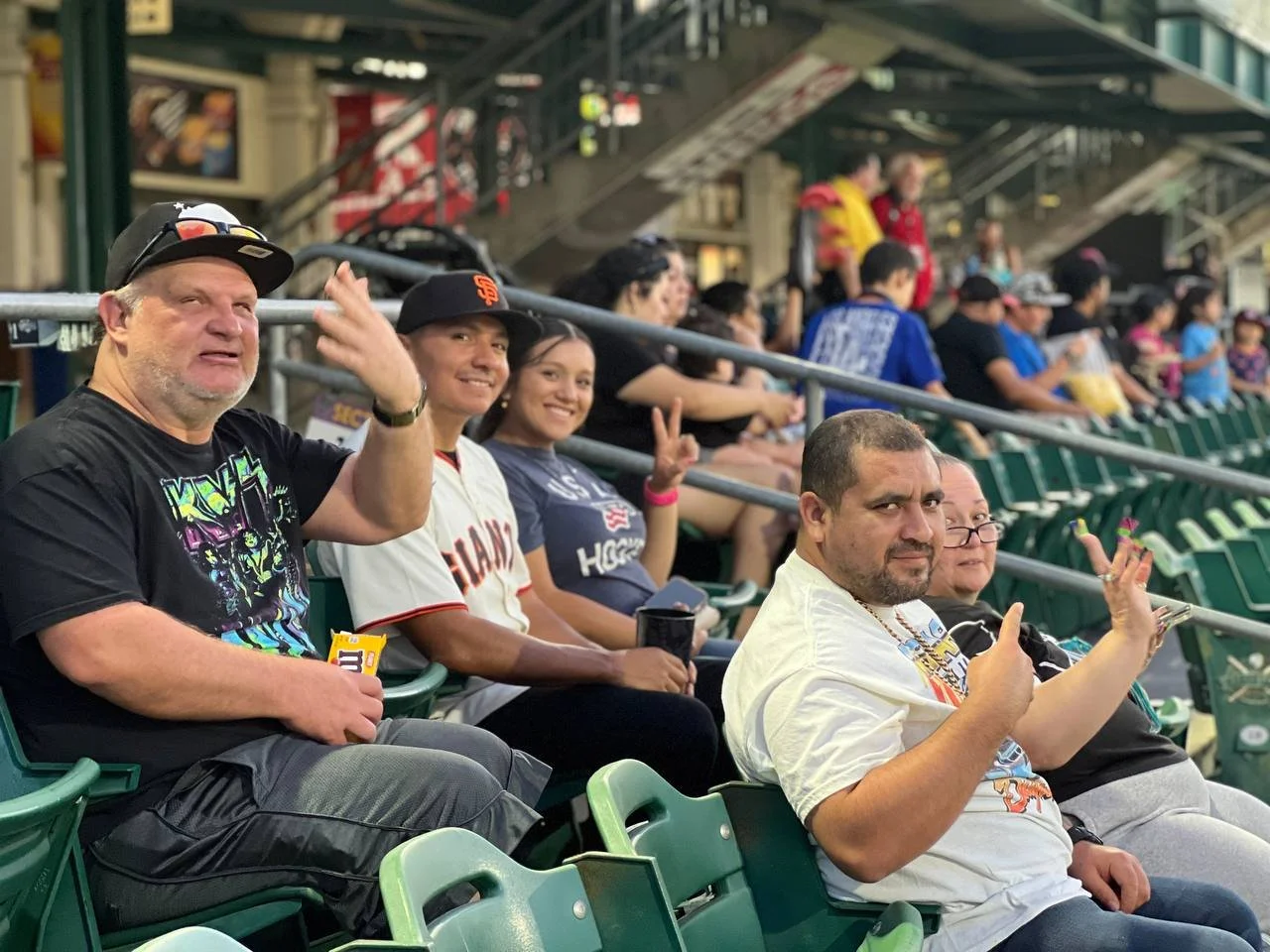 Five smiling people in the stands of a baseball game. Smiling towards the camera with peace-signs up.