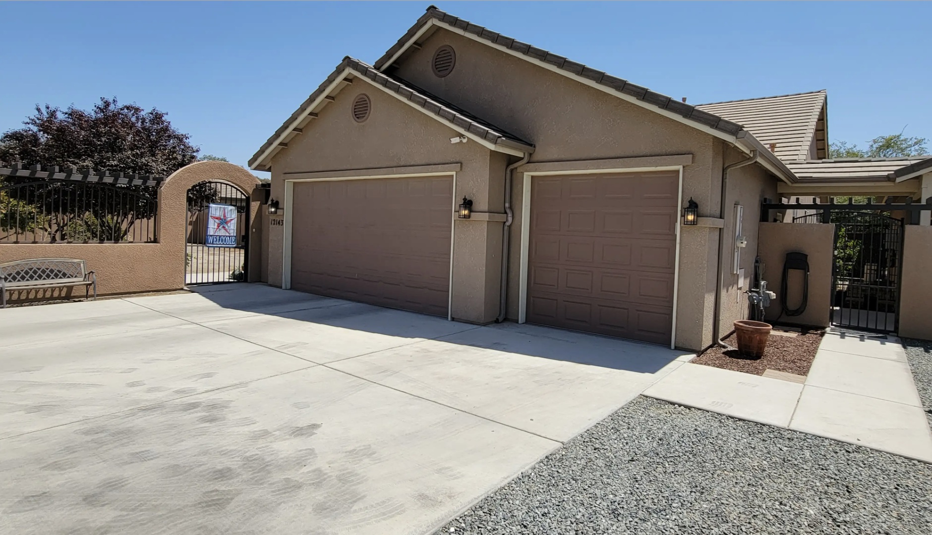 The image shows a residential house with a concrete driveway, two garage doors, and an arched gate with a welcome sign. There is a bench on the left, outdoor wall lights, and a small garden area with a potted plant near the gate.