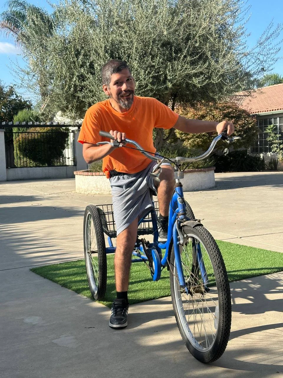 A man with a beard and mustache riding a blue tricycle outdoors on a sunny day, smiling.