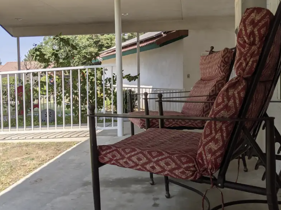 Porch with two cushioned chairs with red patterned upholstery, a white railing, and a view of a garden with trees and shrubs.