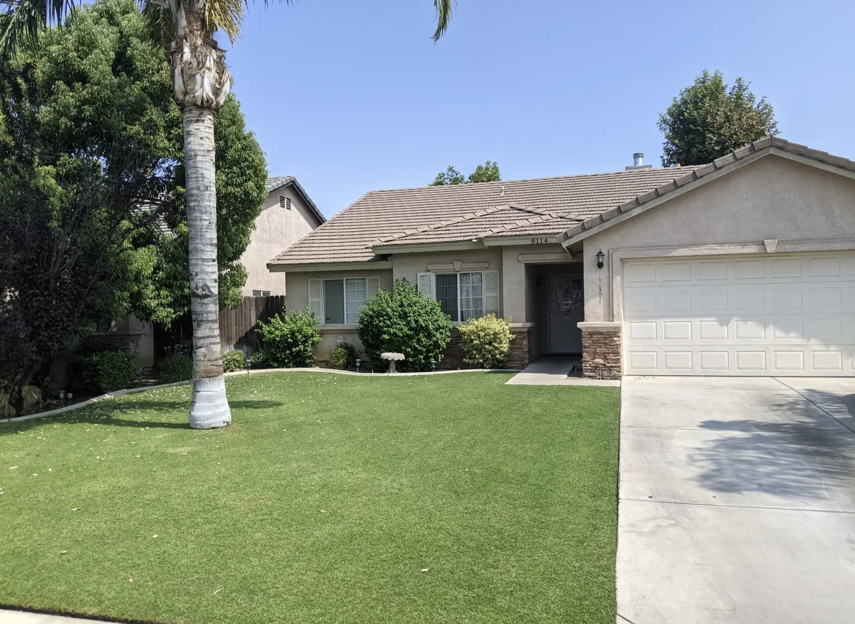Front view of a house with a green lawn, a tall palm tree, bushes, and a driveway leading to a white garage door, under a clear blue sky.
