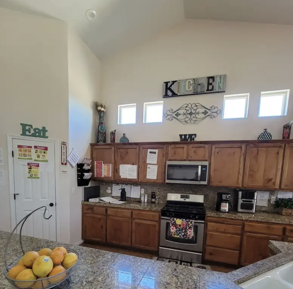 Kitchen with wooden cabinets, granite countertops, a microwave above the stove, a coffee maker, and small decorative items on top of the cabinets. There is a basket of fruit on the counter, and the wall has decorative signs spelling 'KITCHEN' and 'EA