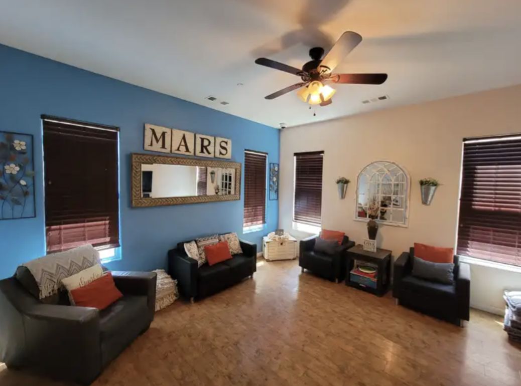 Living room with blue and white walls, four black chairs with red and gray pillows, wooden flooring, and windows with dark blinds. Decor includes a mirror, wall art, and potted plants.