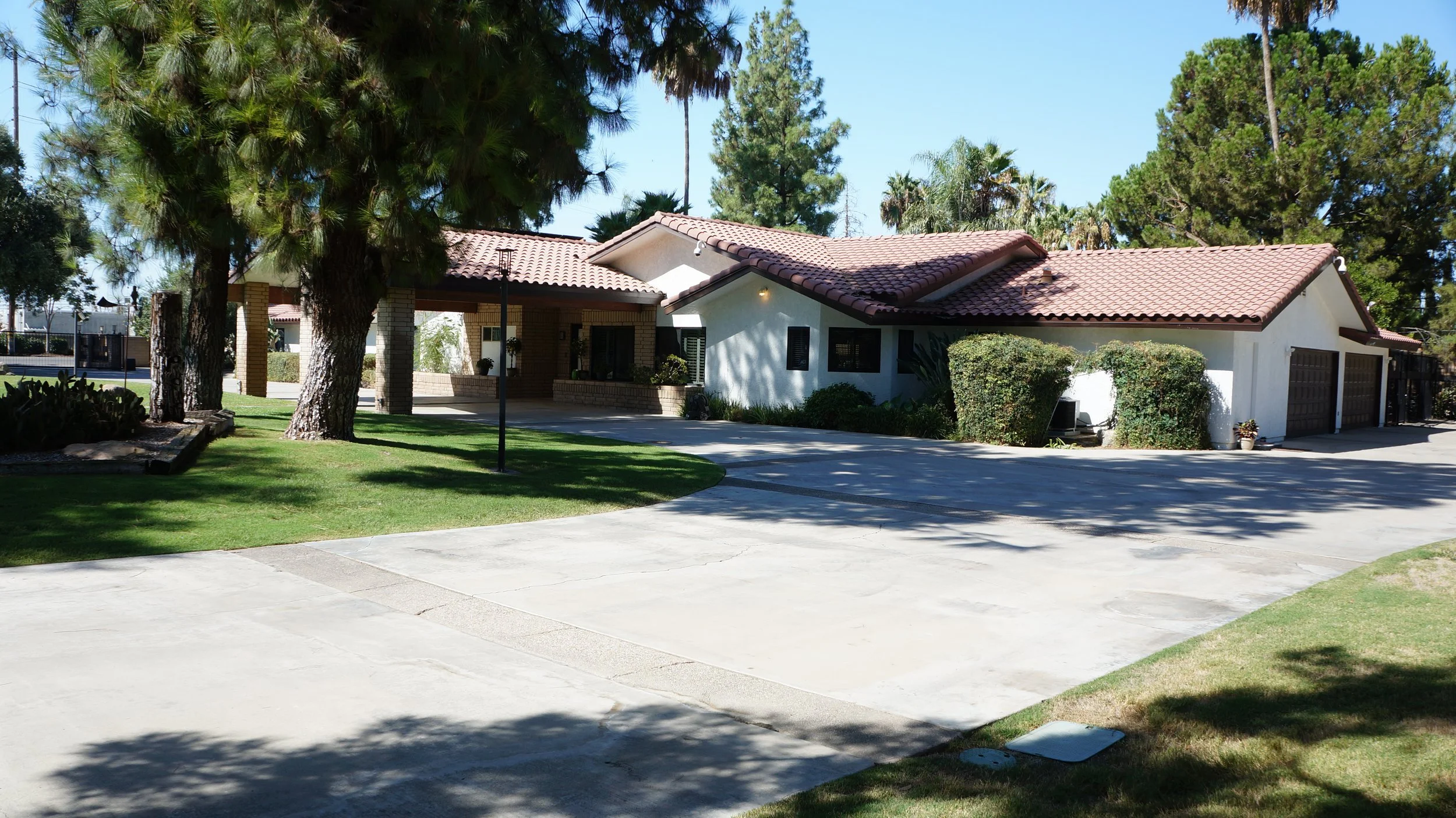 A single-story house with white walls, a red tile roof, and attached garage doors, surrounded by green bushes and trees on a sunny day.