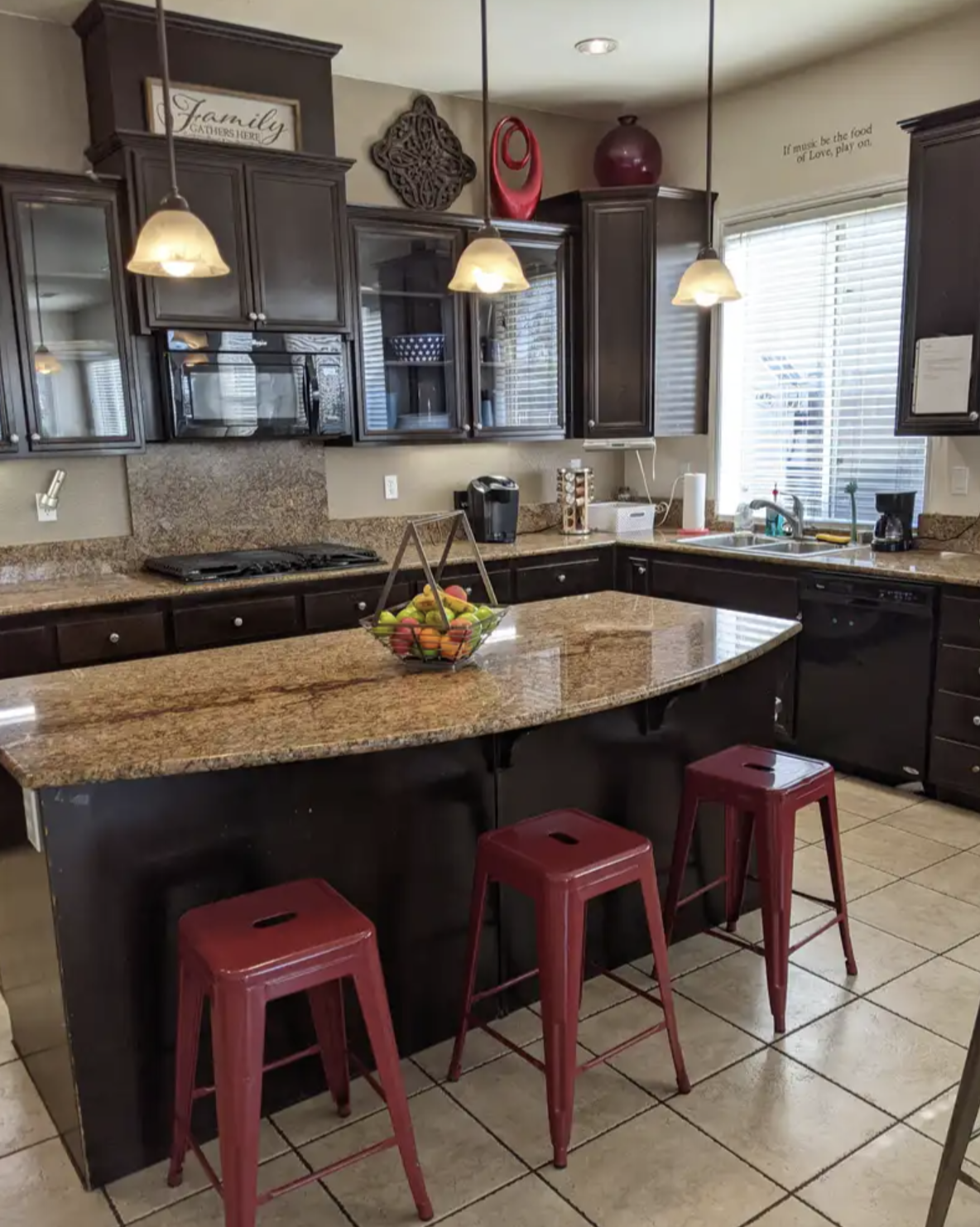 Kitchen with dark wooden cabinets, granite countertops, three red barstools at a kitchen island, and a bowl of fruit, next to a window with blinds.