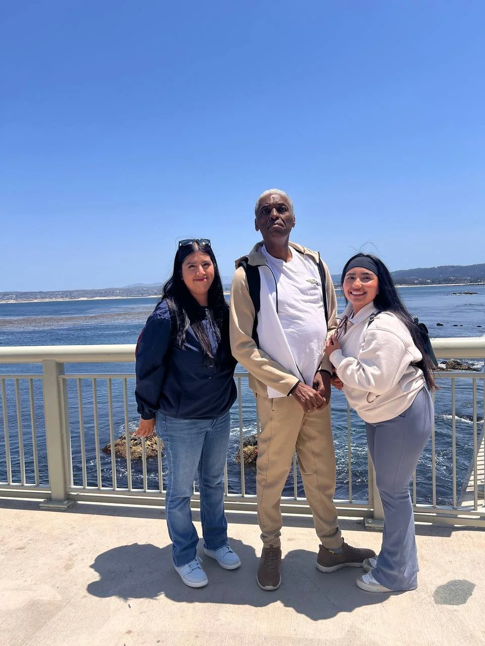 Three people standing on a bridge by the water, with a view of the ocean and distant land under a clear blue sky.